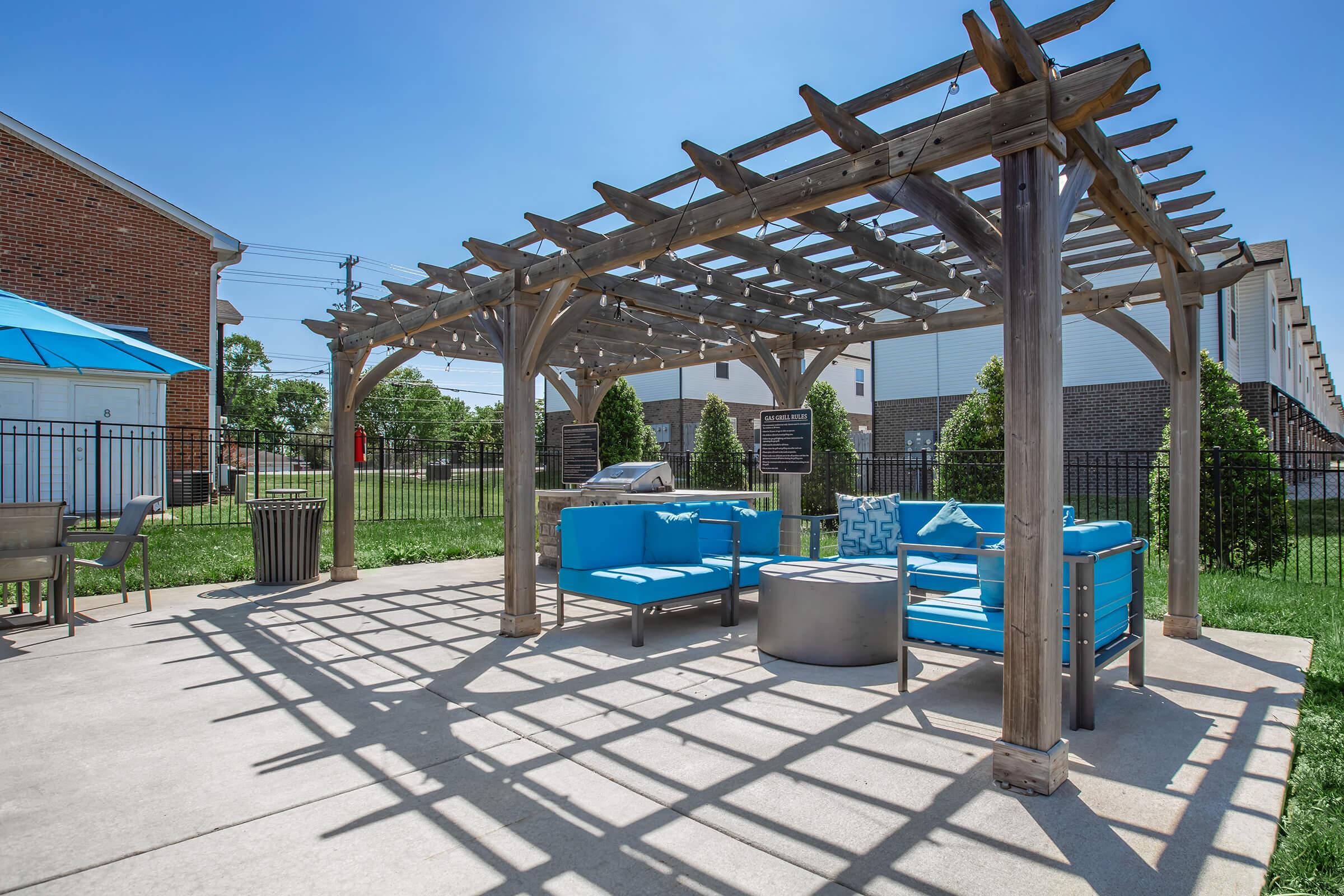 A shaded outdoor seating area featuring a wooden pergola. There are blue lounge chairs, a circular table, and green grass in the background. The area is well-lit with clear skies, creating a relaxing atmosphere.