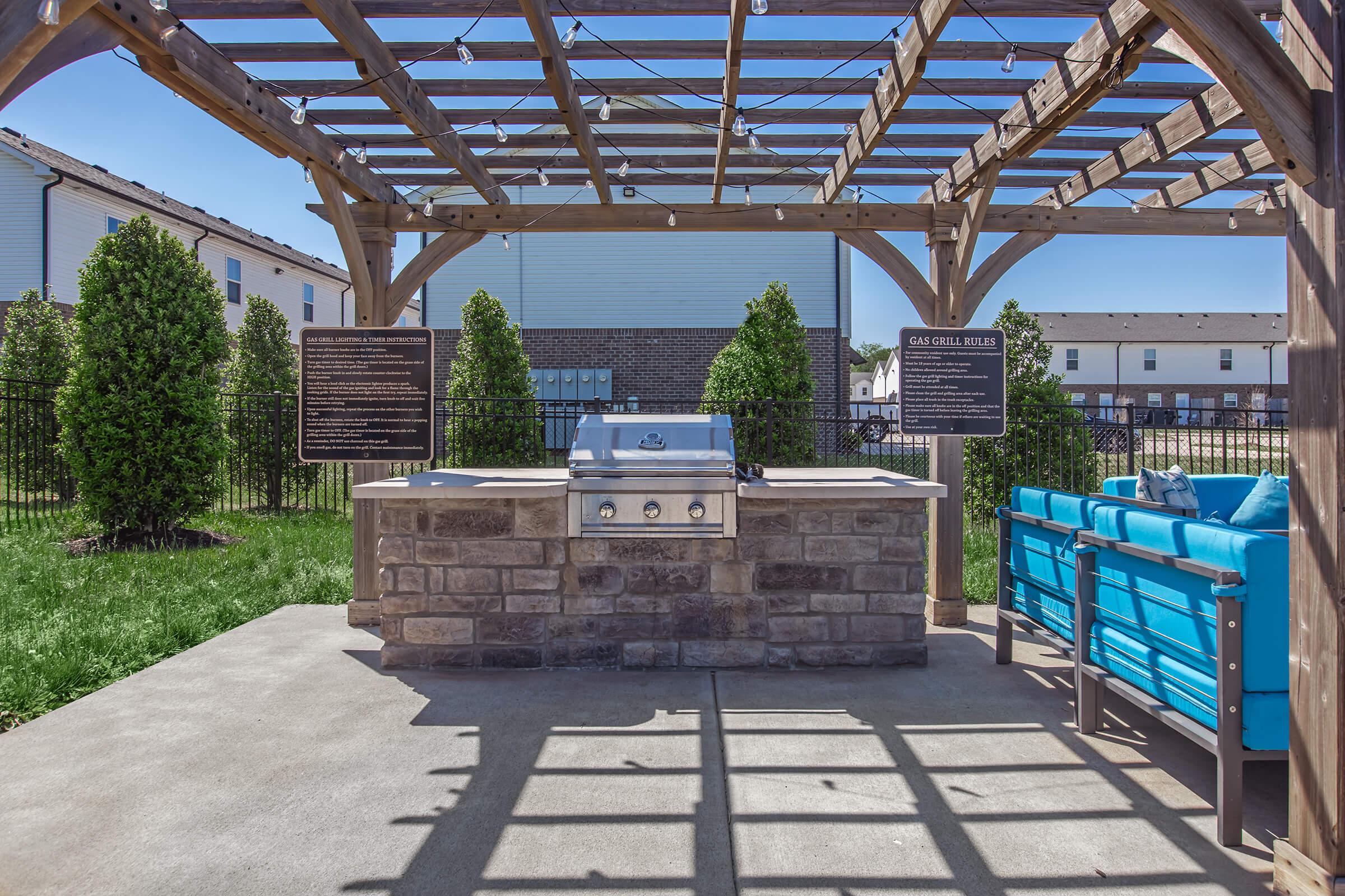 An outdoor grilling area featuring a stone barbecue grill under a wooden pergola. The space includes blue outdoor seating and is surrounded by lush green grass and manicured bushes. Signs are posted on the pergola detailing features of the grill area.