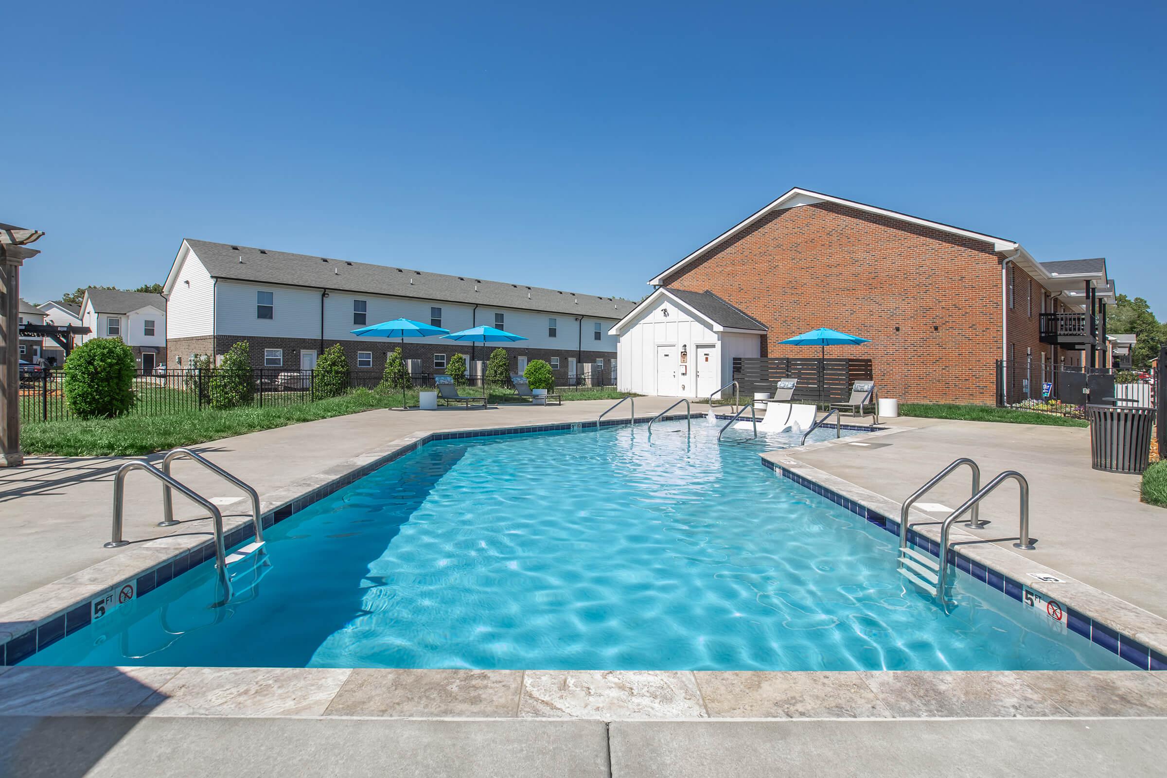 A clear blue swimming pool surrounded by lounge chairs and umbrellas. In the background, there are several residential buildings and a well-maintained grassy area. The sky is bright and clear, indicating a sunny day. The pool area is inviting and well-designed for relaxation and leisure activities.