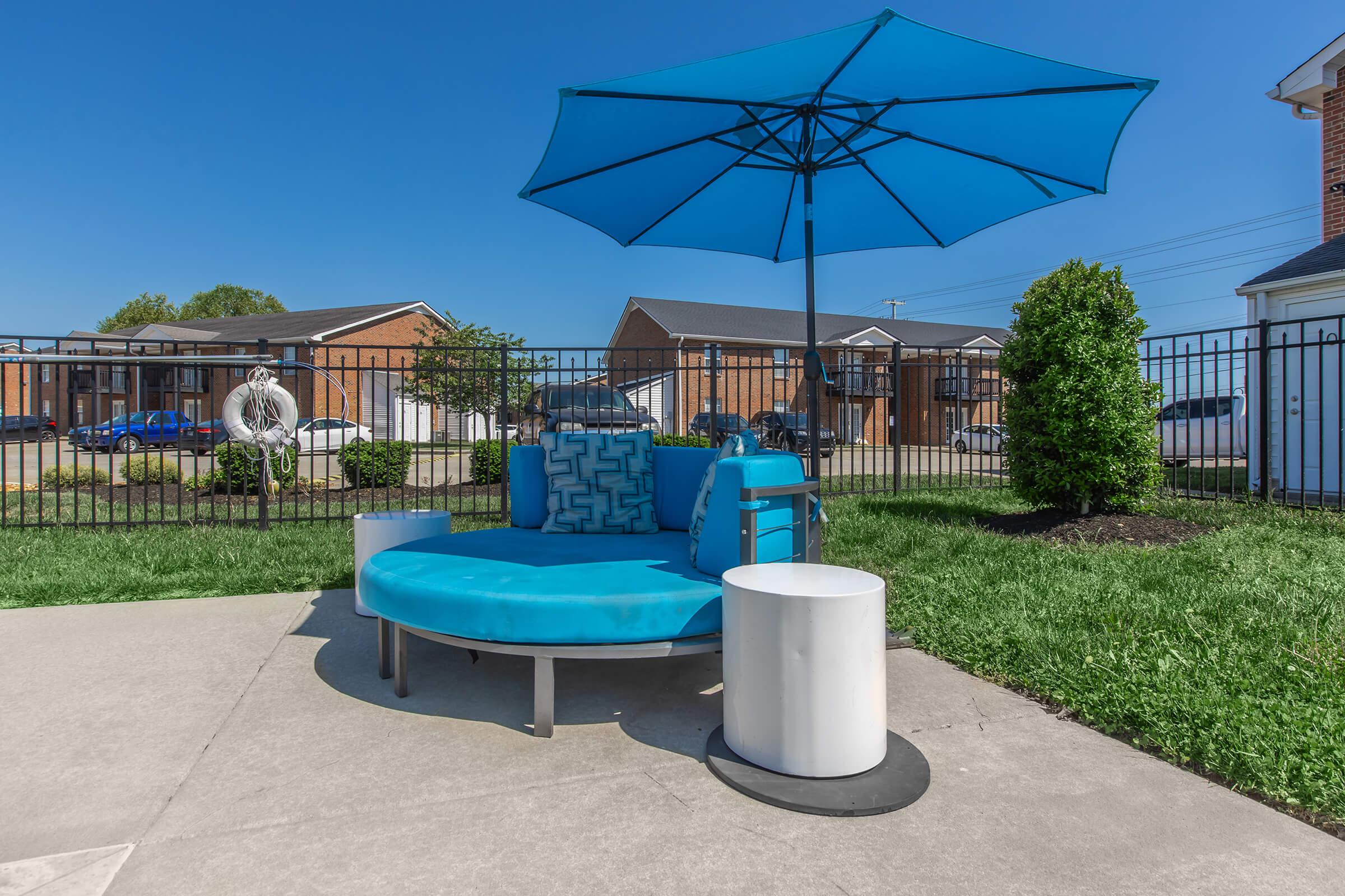 A circular blue lounge chair with an umbrella, accompanied by two small side tables. The setting features a well-maintained lawn and a fence in the background, with a few buildings and parked cars visible. The clear blue sky adds to the outdoor ambiance.