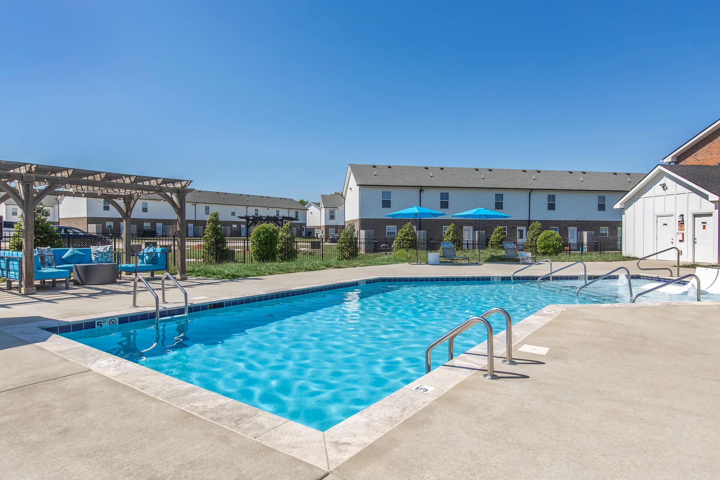 A swimming pool with clear blue water, surrounded by a paved area. There are lounge chairs, umbrellas, and a nearby pergola. In the background, there are several residential buildings with a clear blue sky overhead.