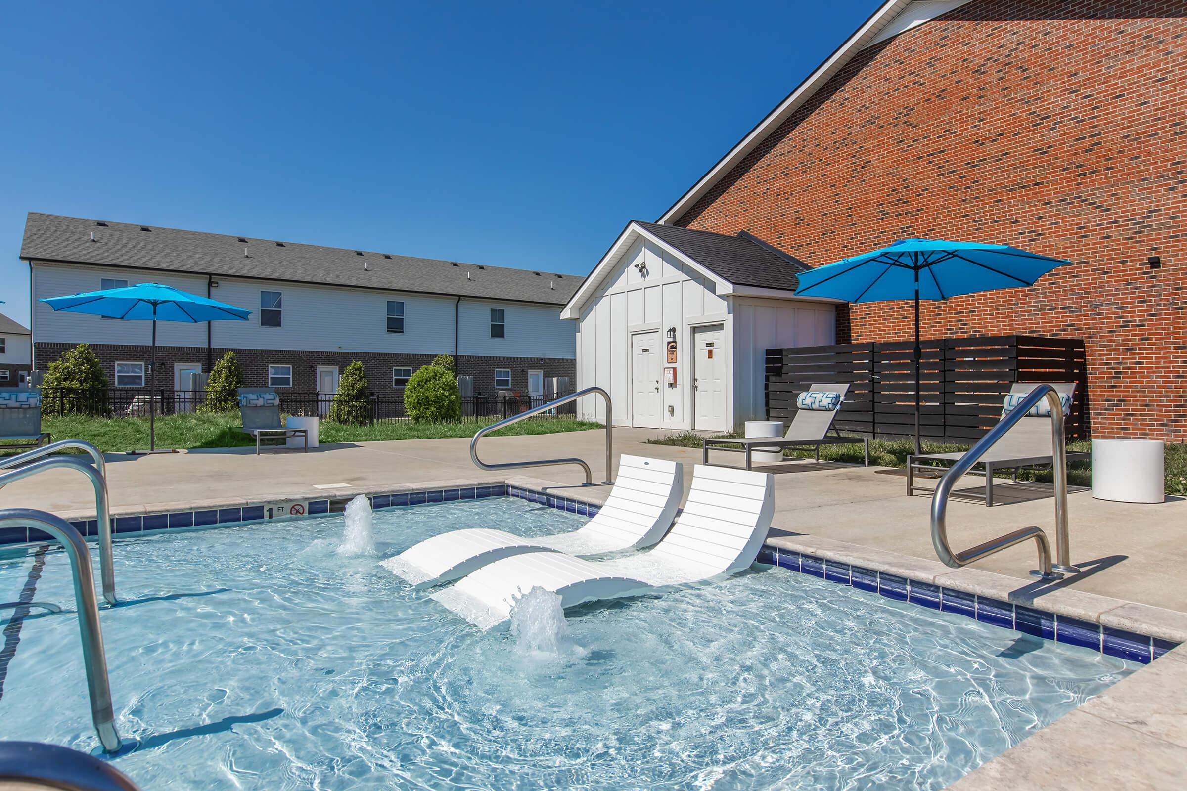A swimming pool area featuring a white lounge chair partially submerged in water, with a splash feature. Surrounding the pool are blue umbrellas, lounge seating, and a white maintenance building in the background. The scene is set under a clear blue sky, with grassy areas visible.