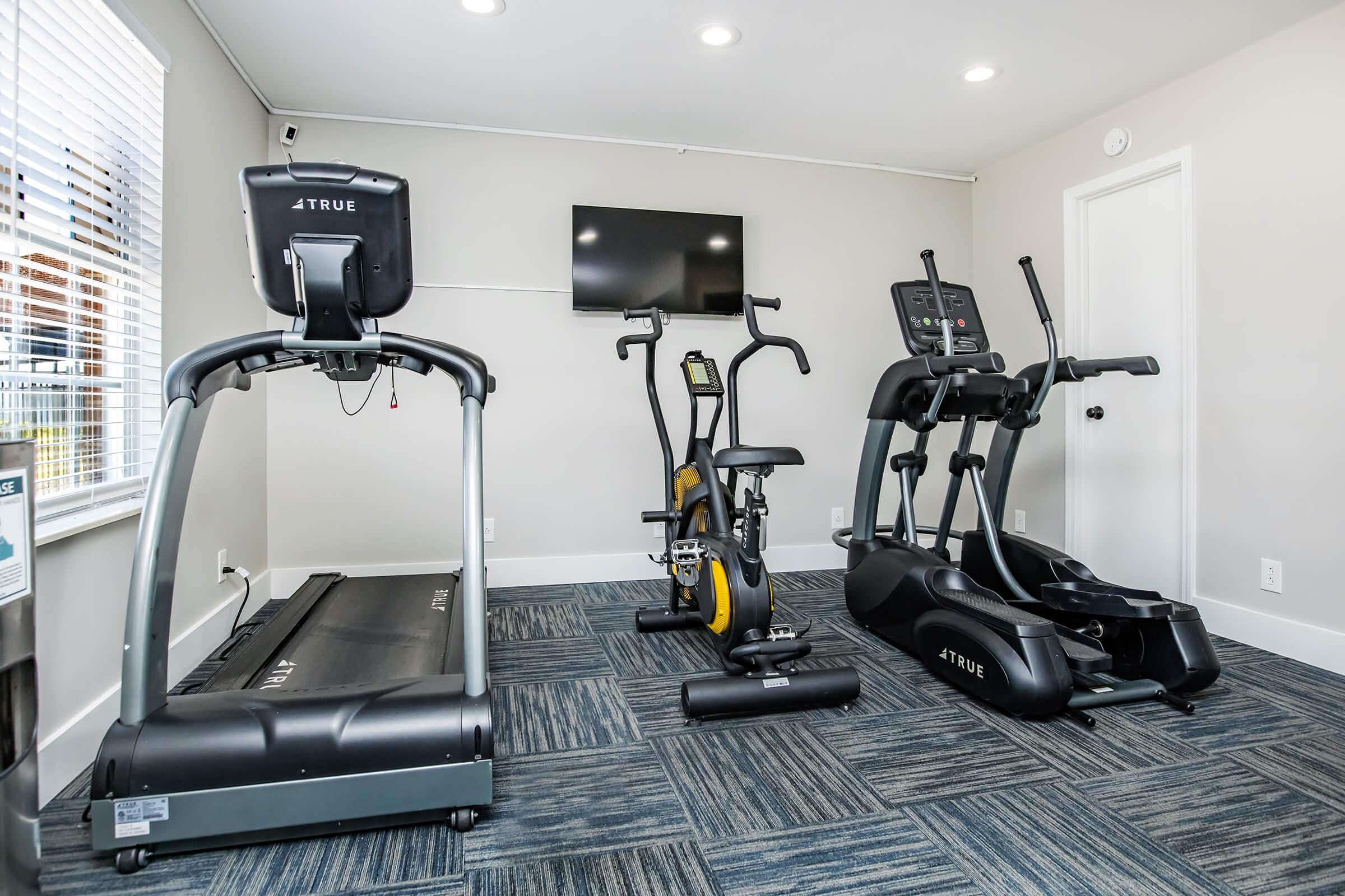 A modern fitness room featuring a treadmill, stationary bike, and elliptical machine, all in sleek black and yellow. The room has bright lighting, a large window, and a wall-mounted television, creating a welcoming environment for exercise. The floor is covered with a blue carpet.