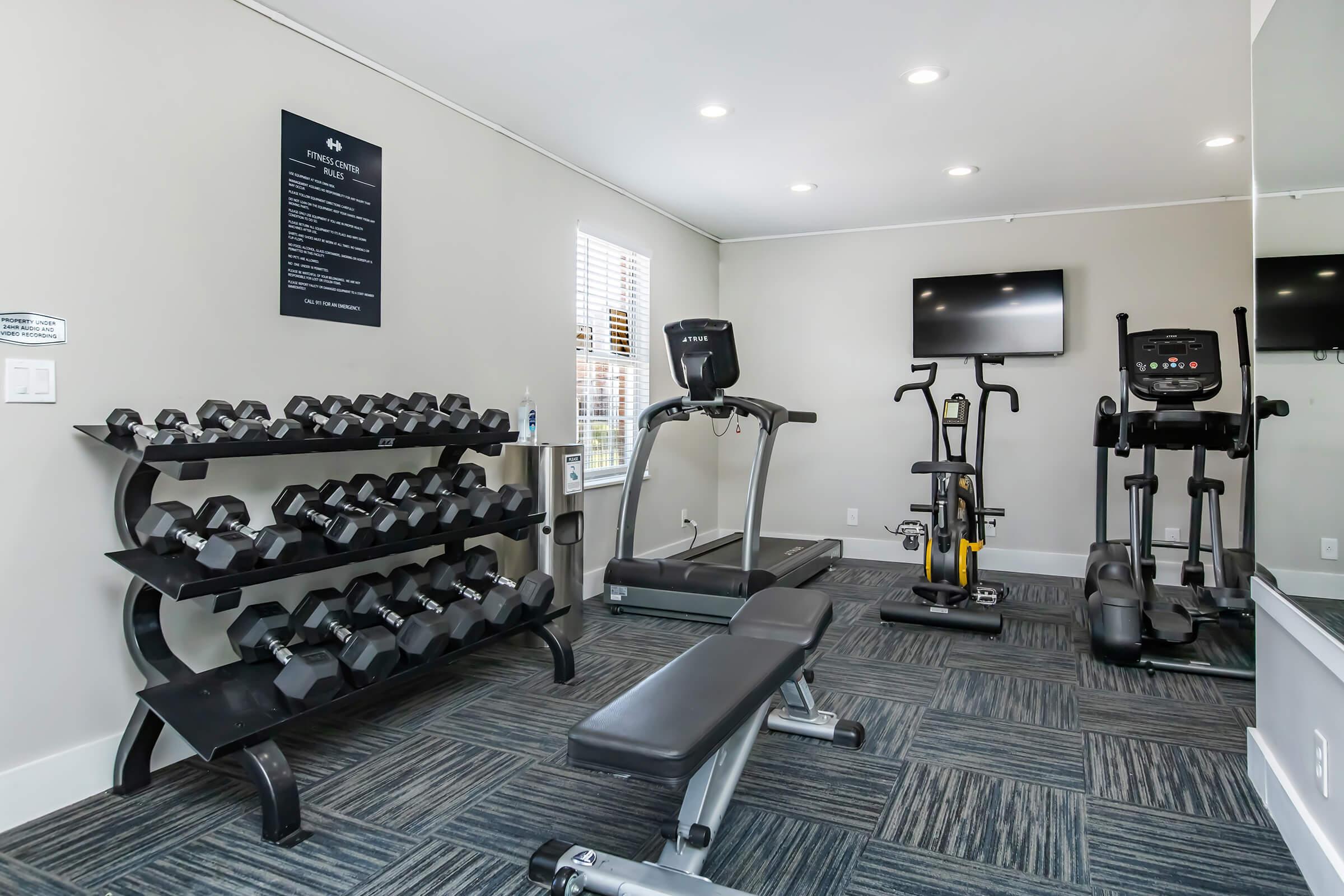 A modern gym interior featuring a rack of dumbbells, a treadmill, an elliptical machine, and a stationary bike. The space has bright lighting, a flat-screen TV mounted on the wall, and a mirror, with grey and blue carpeting on the floor. Windows allow natural light in.