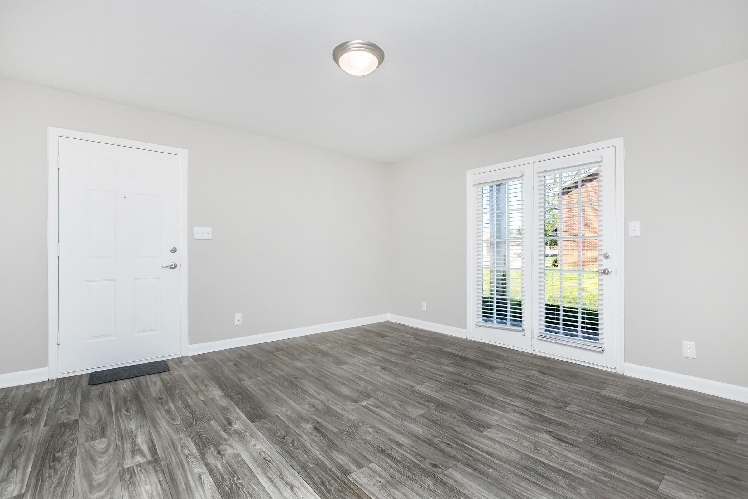 An empty room featuring light gray walls, a wooden floor, and a ceiling light. The room has a front door and a set of glass doors with white shutters leading to an outdoor area. Natural light comes through the doors, creating a bright and airy atmosphere.