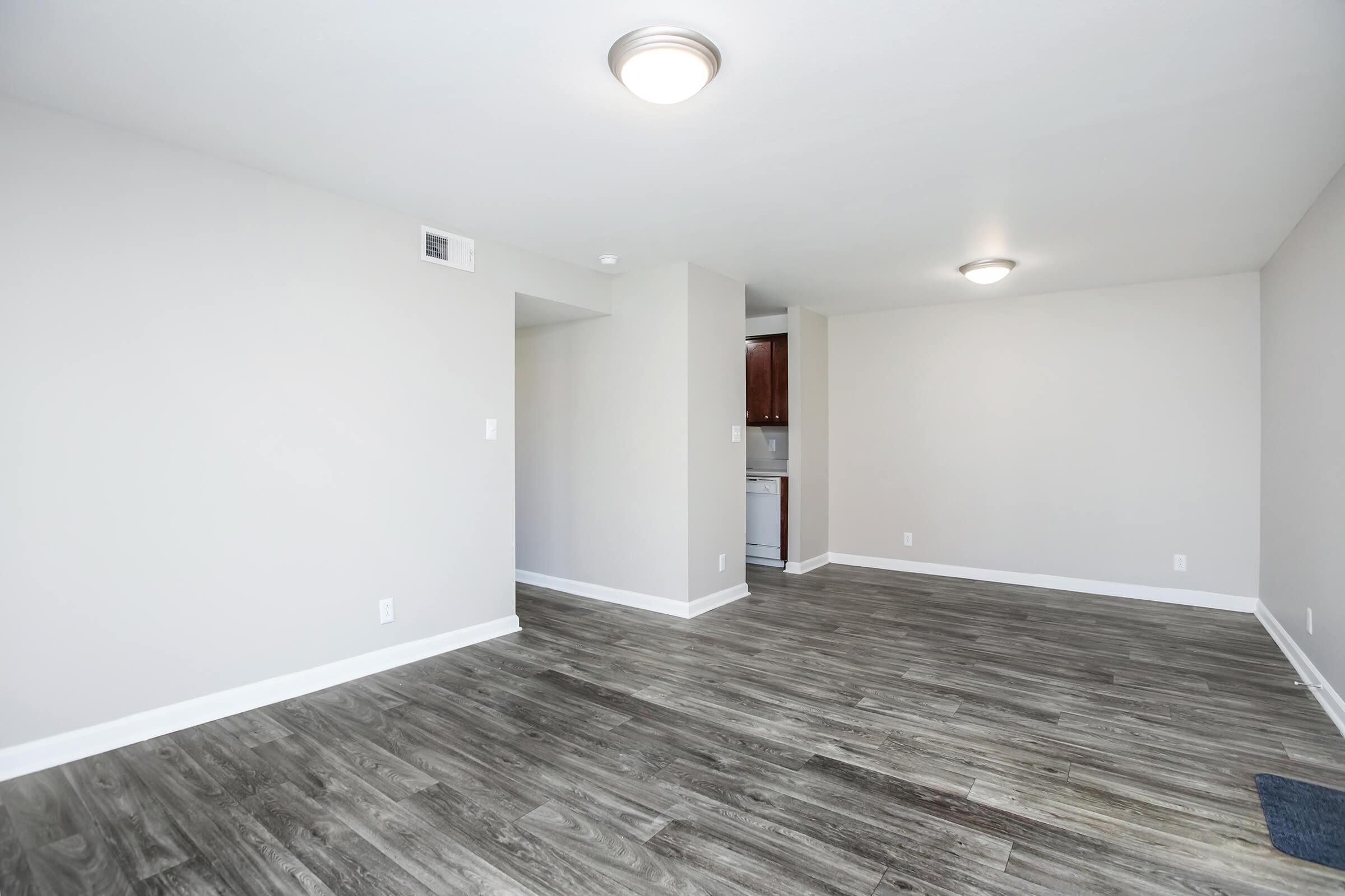 A spacious, empty room with light gray walls and a hardwood floor. The ceiling features a round light fixture. In the background, there's a glimpse of a kitchen area with wooden cabinets. The overall atmosphere is bright and modern, providing a blank canvas for decoration.