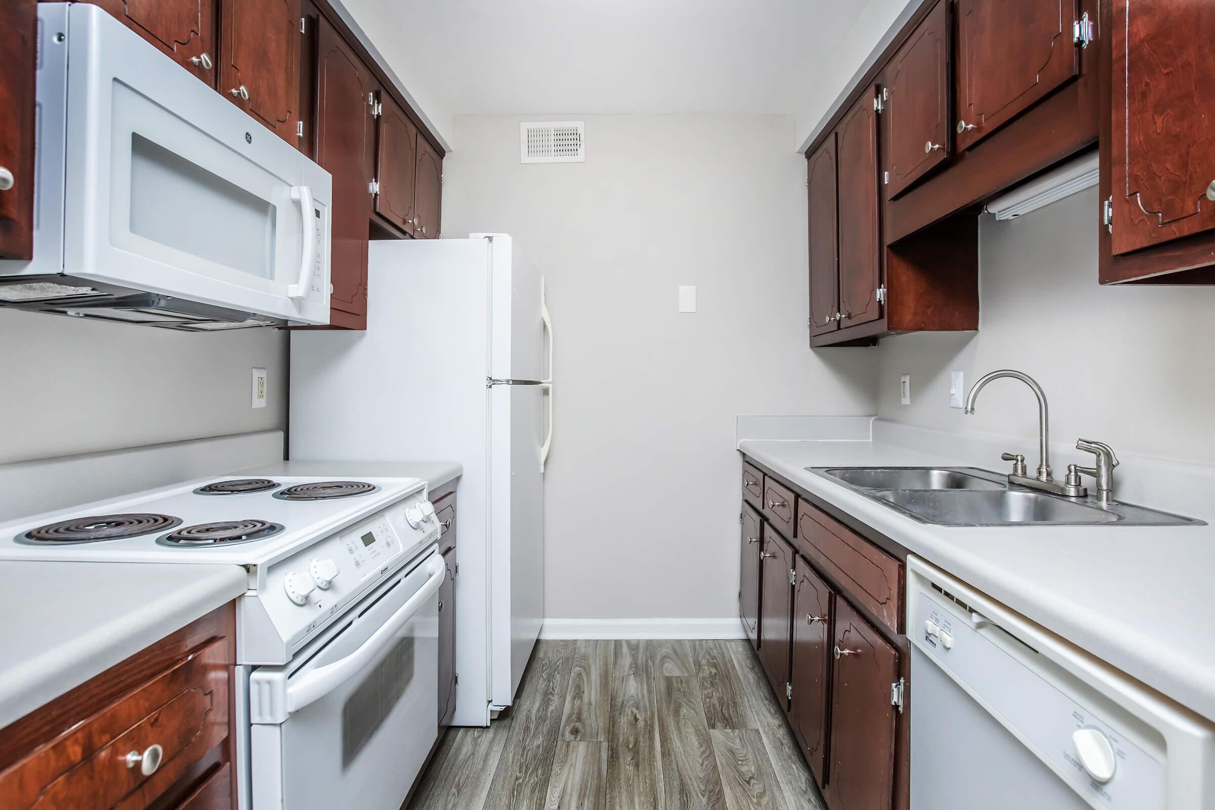 A modern kitchen featuring dark wood cabinets, a white refrigerator, a white microwave, an oven with stovetop, and a stainless steel sink. The countertops are light-colored, and the flooring is wood-look laminate. The walls are painted a soft gray, creating a bright and inviting atmosphere.