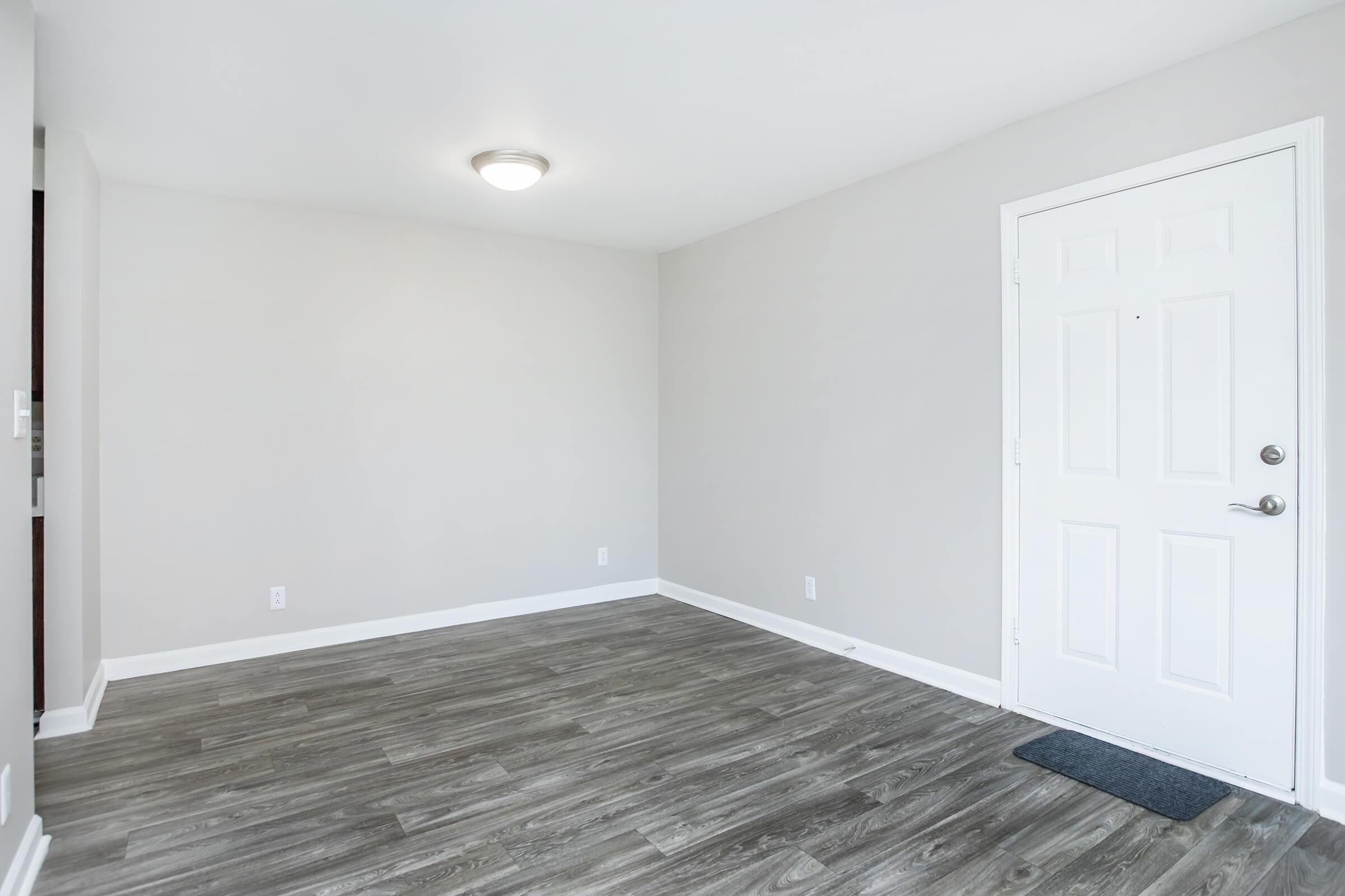 Empty room with light gray walls and a white door. The flooring is light wood laminate. A ceiling light fixture is visible. There are no furniture or decorations in the space, creating a clean and minimalist appearance.