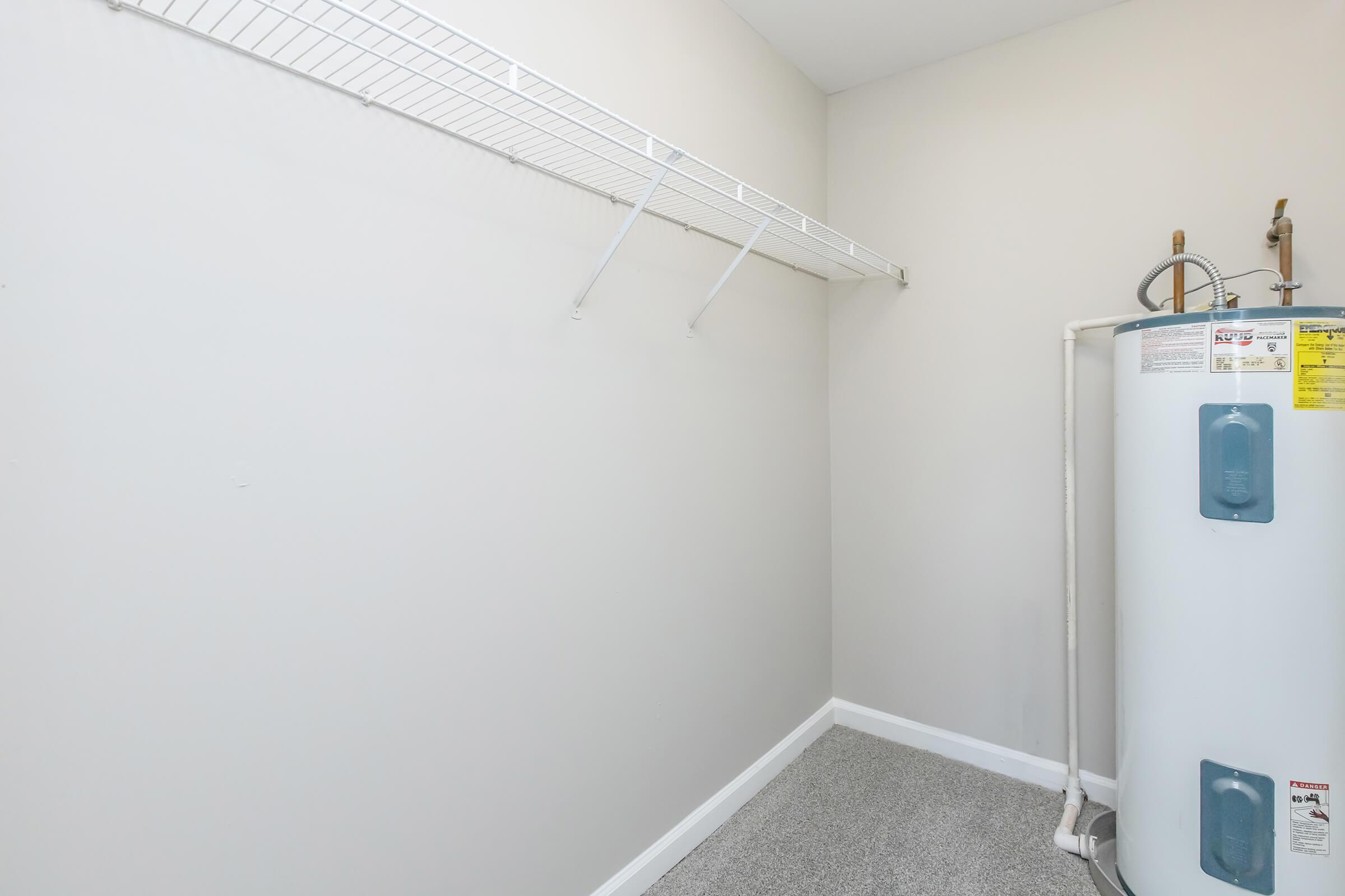 A small, empty utility room featuring a white wall, a wire shelf mounted on the wall, and a water heater on the right side. The floor is covered with light gray carpet, creating a minimalist and functional space.