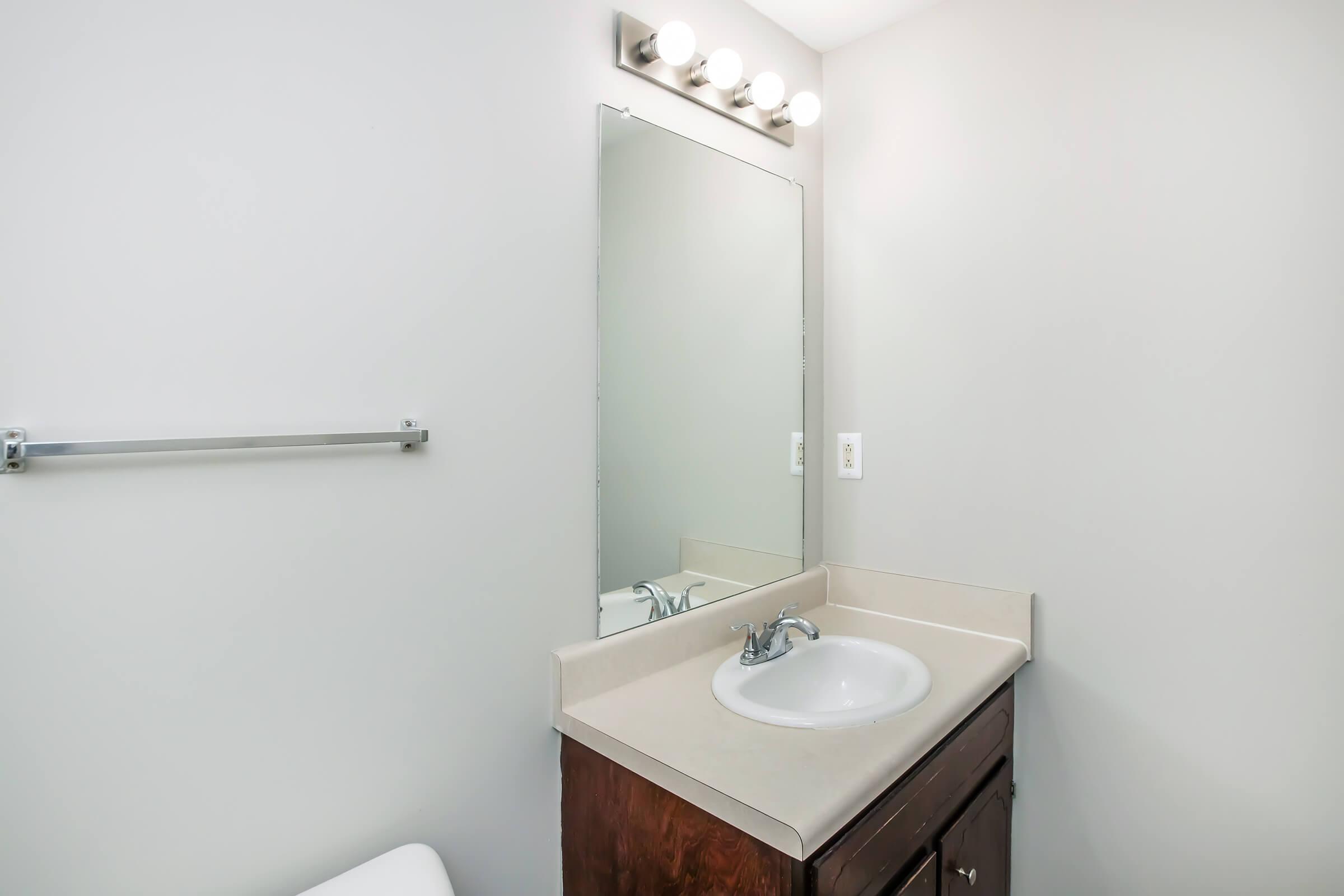 A simple bathroom featuring a beige countertop with a sink, a framed mirror above it, and a strip of bright lights mounted on the wall. A towel rack is positioned on the left, and a white toilet is partially visible in the foreground. The walls are painted a light gray.