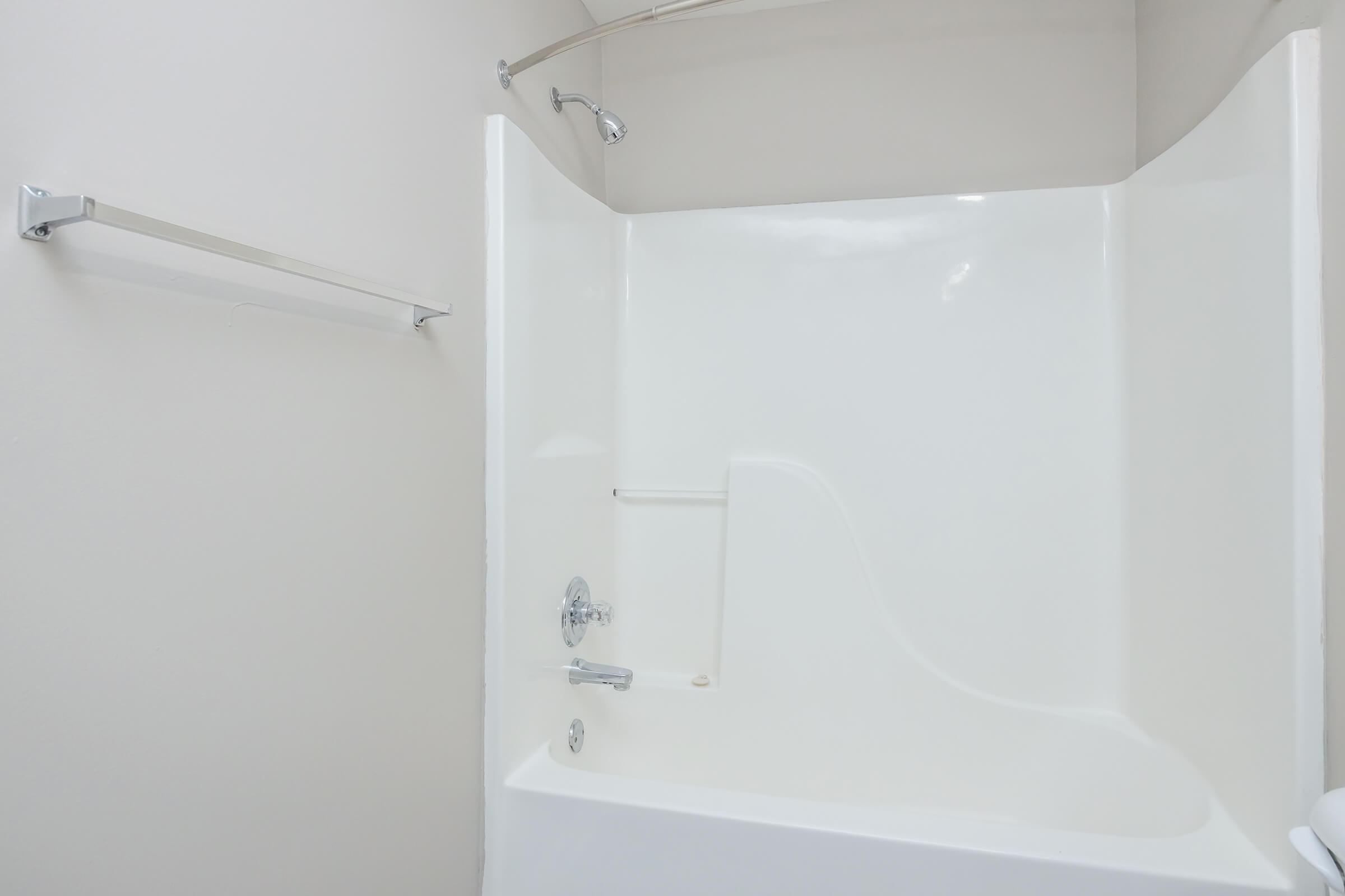 A clean, white bathtub with a showerhead and a modern design. The walls are smooth and light-colored, and there is a chrome towel bar mounted on the side. The space has a minimalistic aesthetic with no visible decorations or personal items.