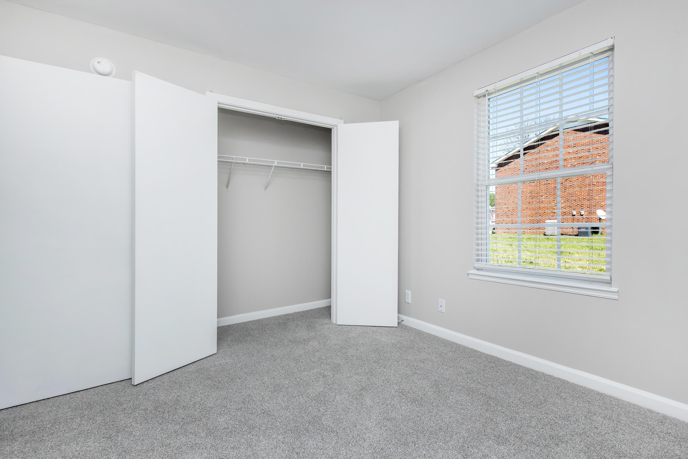 A light-colored room with beige walls and grey carpet. It features an open white closet with no items inside and a window with white blinds, allowing natural light to enter. Outside the window, a glimpse of a brick building can be seen.