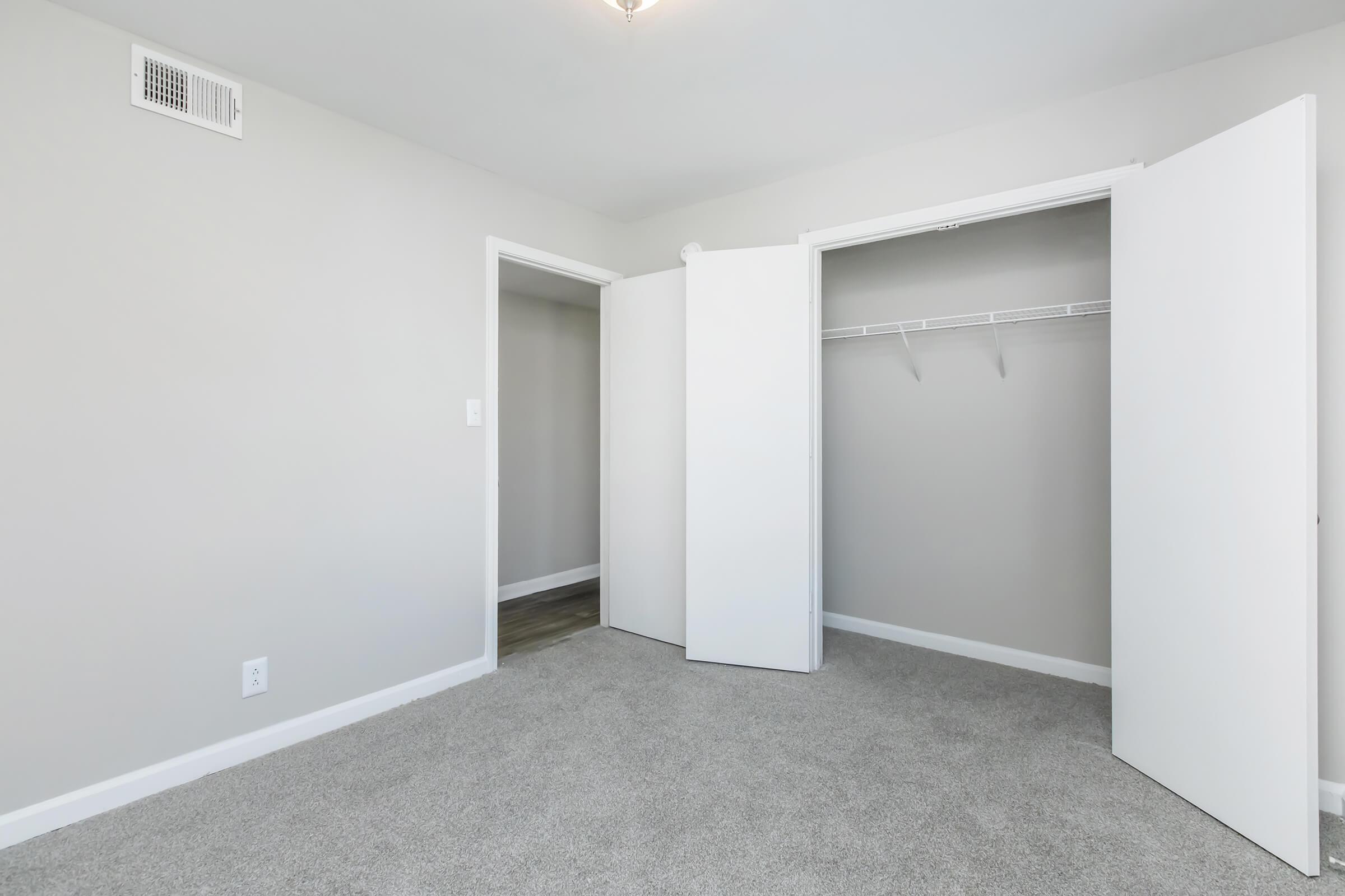 A simple, empty room with light gray walls and carpeted flooring. Two open closet doors on the right reveal a hanging rod inside. There are two doorways leading to other areas, one to the left and another straight ahead. A ceiling light illuminates the space.