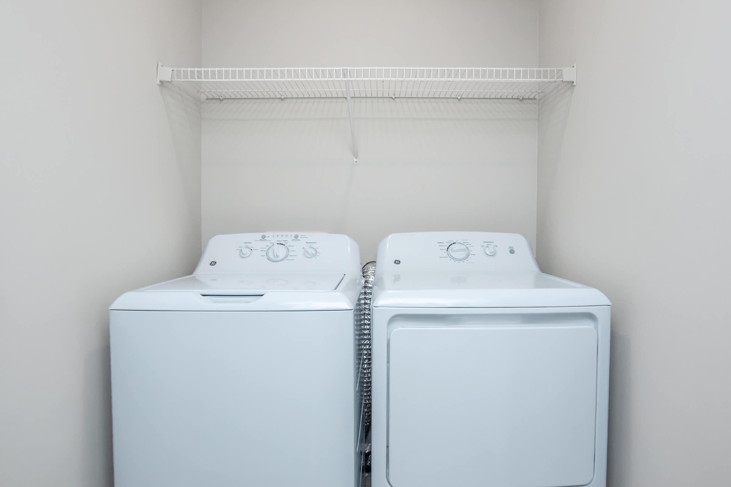 A clean laundry room featuring a white washing machine and a white dryer side by side, set against a light-colored wall. Above them, there is a simple wire shelf for storage. The overall aesthetic is modern and minimalist.