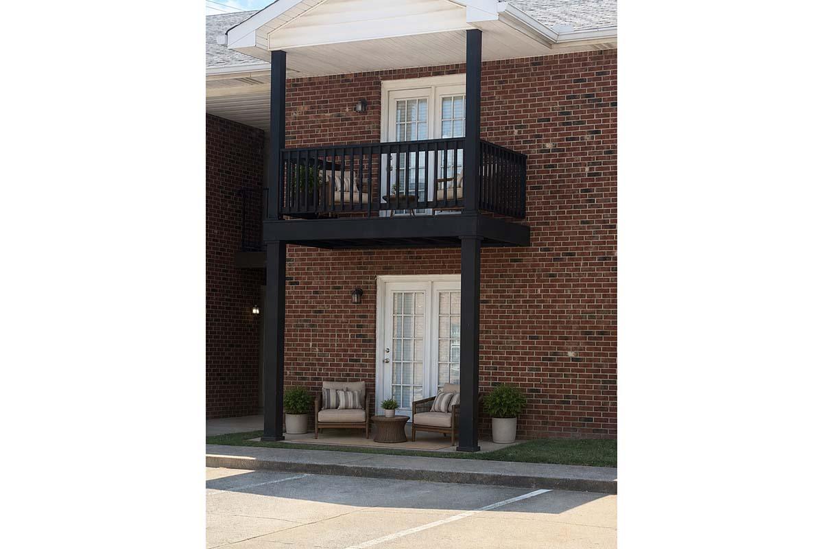A two-story brick building with a balcony above. The balcony has a black railing and two chairs. Below, there are two potted plants beside the seating area. The exterior features a door with large windows, and a parking lot is visible in the foreground.