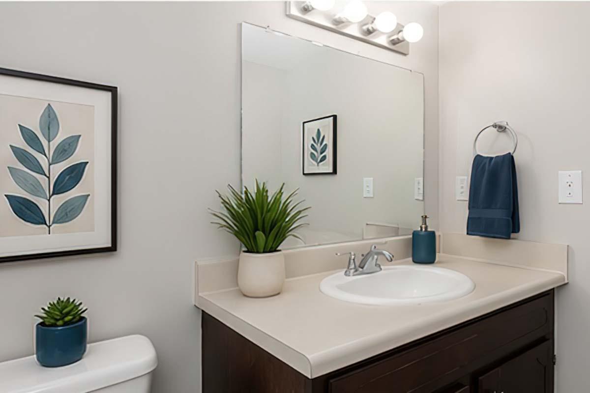 A modern bathroom featuring a light-colored countertop with a sink, a mirror above it, and a hand towel on a towel rack. Decorative elements include a framed leaf print on the wall, a green plant in a pot, and a small succulent on the counter. Soft, neutral color scheme creates a calming atmosphere.