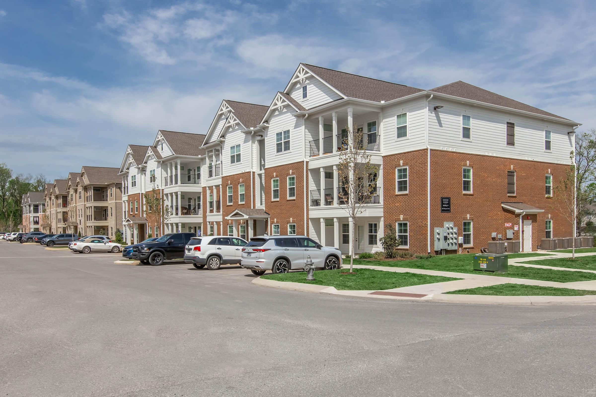 A row of modern multi-family apartment buildings featuring a mix of brick and siding exteriors. The scene includes parked cars along the street, manicured grass, and a clear blue sky. The architecture displays a suburban residential aesthetic with balconies and well-maintained landscaping.