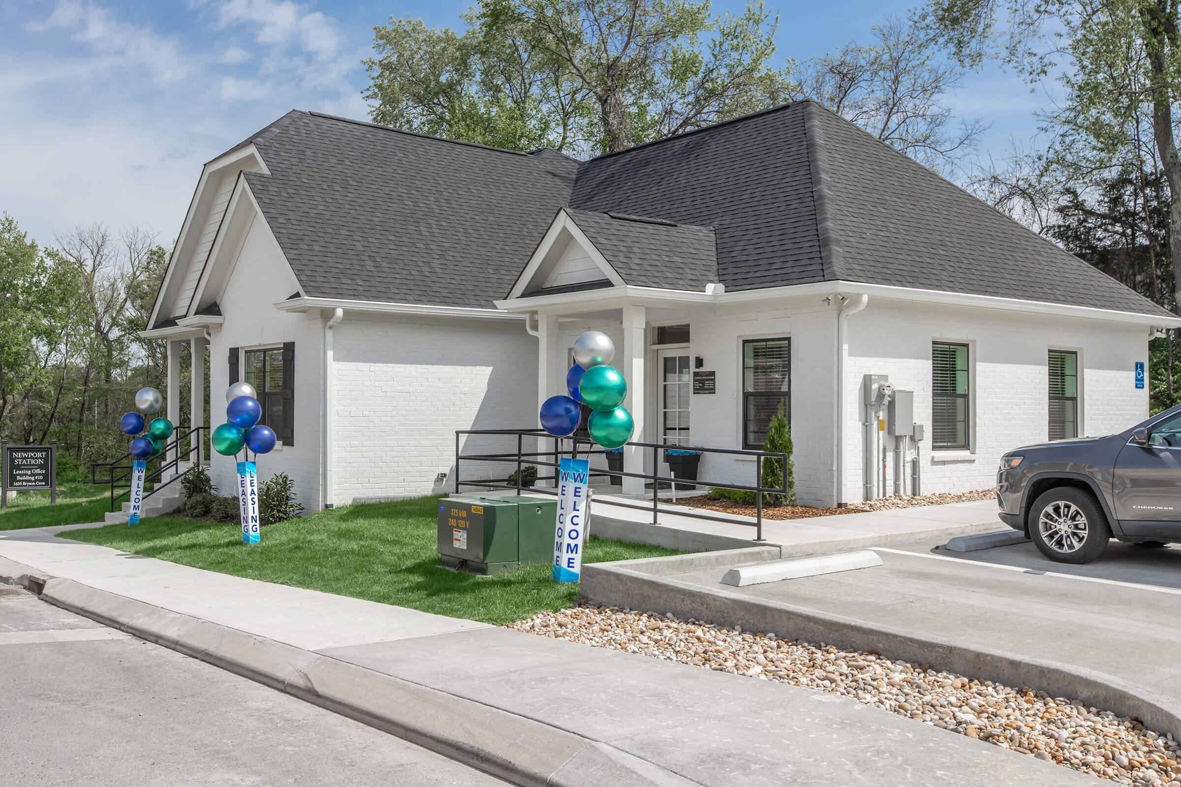 A modern white building with a black roof, featuring large windows. The entrance is flanked by blue and green balloons on tall stands. A sign reads "Welcome." There are landscaped areas with small shrubs and a parking space in front, with a gray vehicle parked nearby. The sky is clear with a few clouds.
