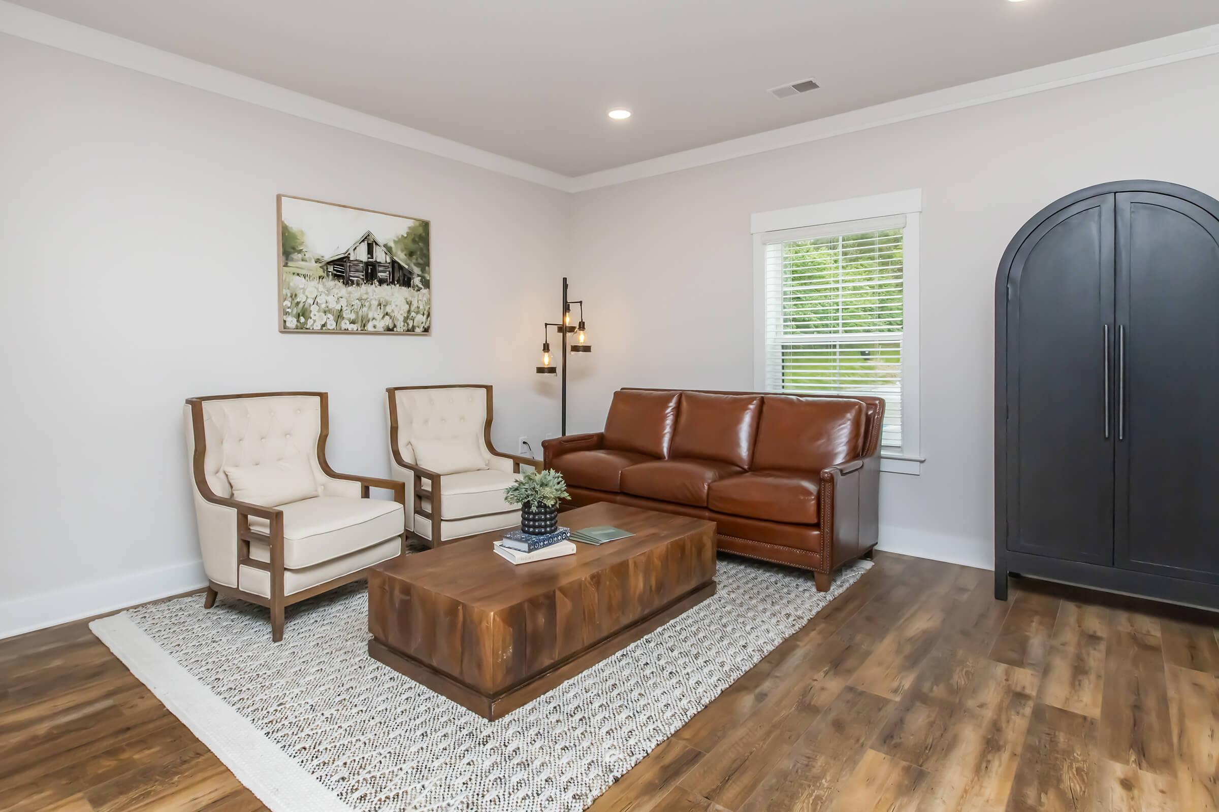 A modern living room featuring a brown leather sofa, two cream-colored upholstered chairs, and a wooden coffee table on a textured rug. The walls are painted light, and there's a framed landscape painting. Natural light streams through a window, creating a warm and inviting atmosphere.