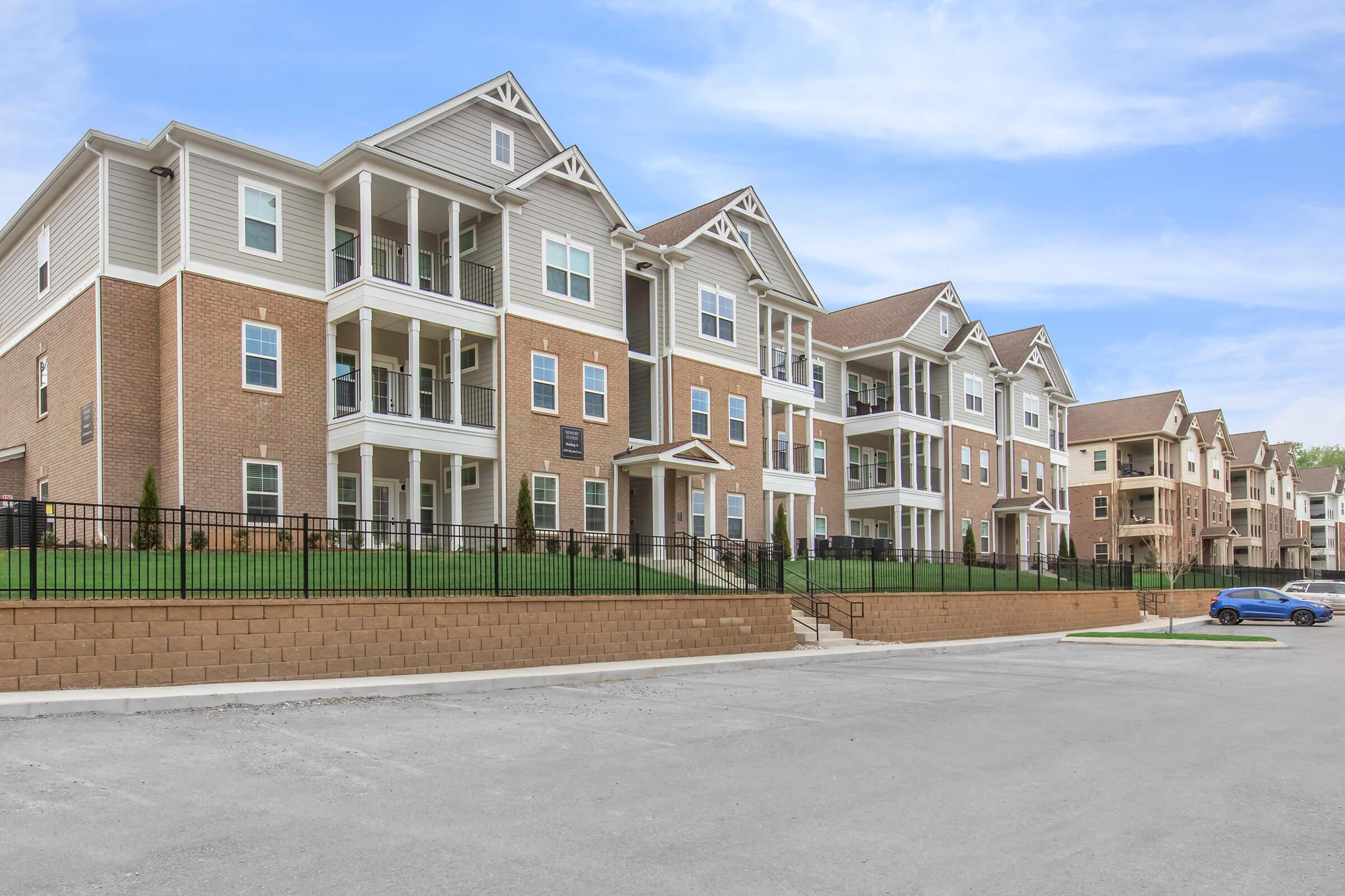 A modern multi-story apartment building featuring a mix of brick and siding exterior. The building has multiple balconies, surrounded by green grass and a landscaped area. In the foreground, there is a paved parking area with a blue car parked. The sky is clear and blue, suggesting a sunny day.