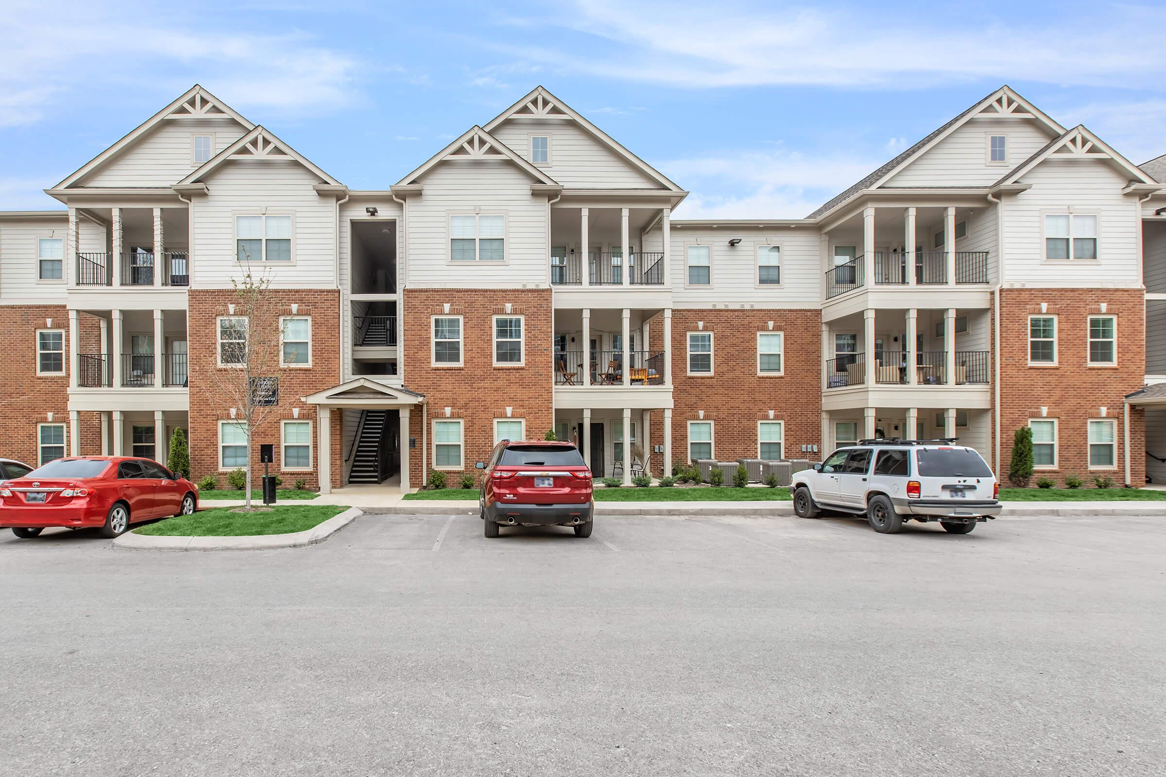 A three-story apartment building with a mix of brick and siding façade. The building features multiple balconies and staircases. In the foreground, there are several parked cars, including two red vehicles and a white one, with a landscaped area and walkways visible. The sky is blue with a few clouds.