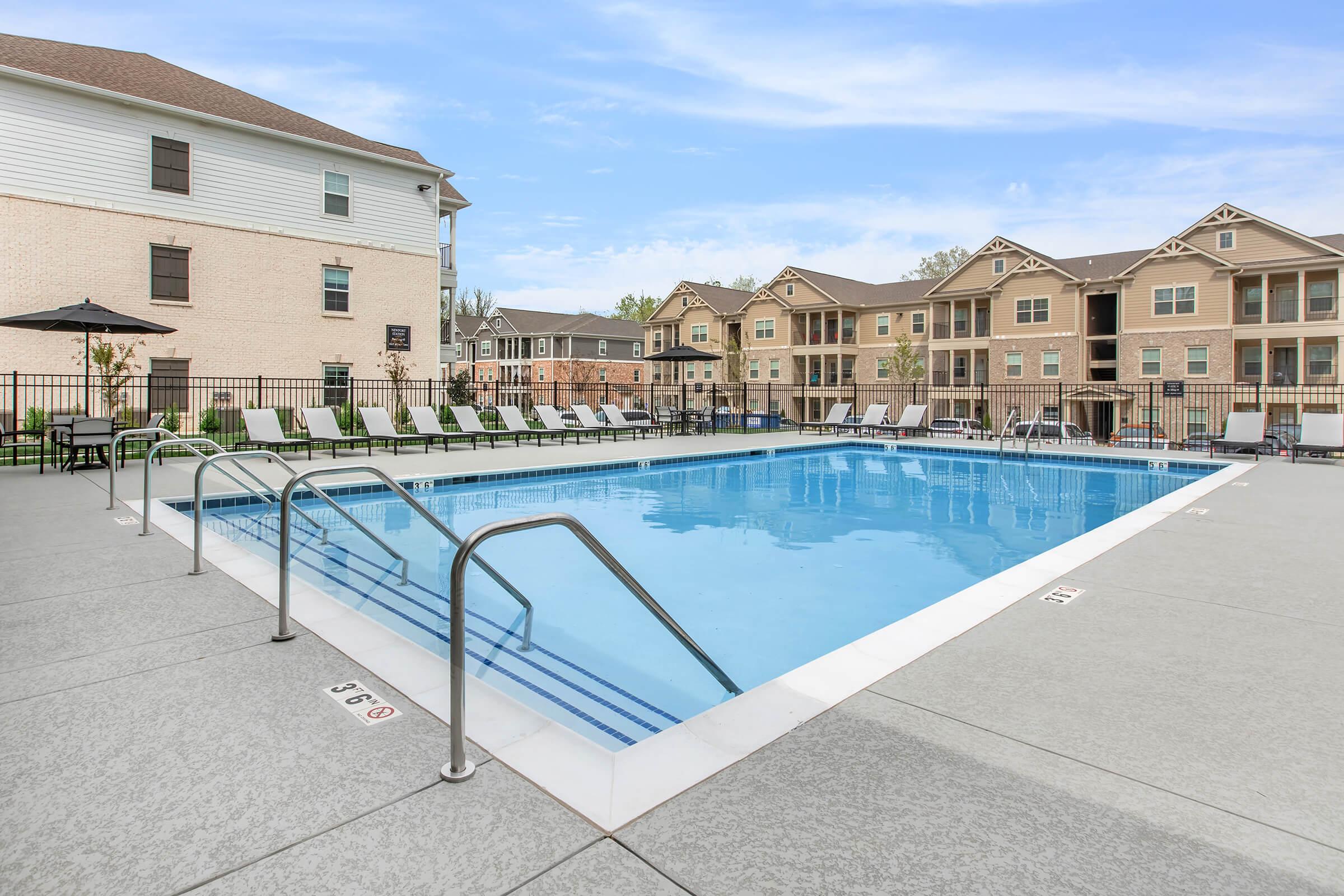 A clean swimming pool area with steps leading into the water, surrounded by lounge chairs. In the background, several multi-story residential buildings are visible, under a clear blue sky. The pool area is well-maintained, with a few tables and umbrellas for shade.