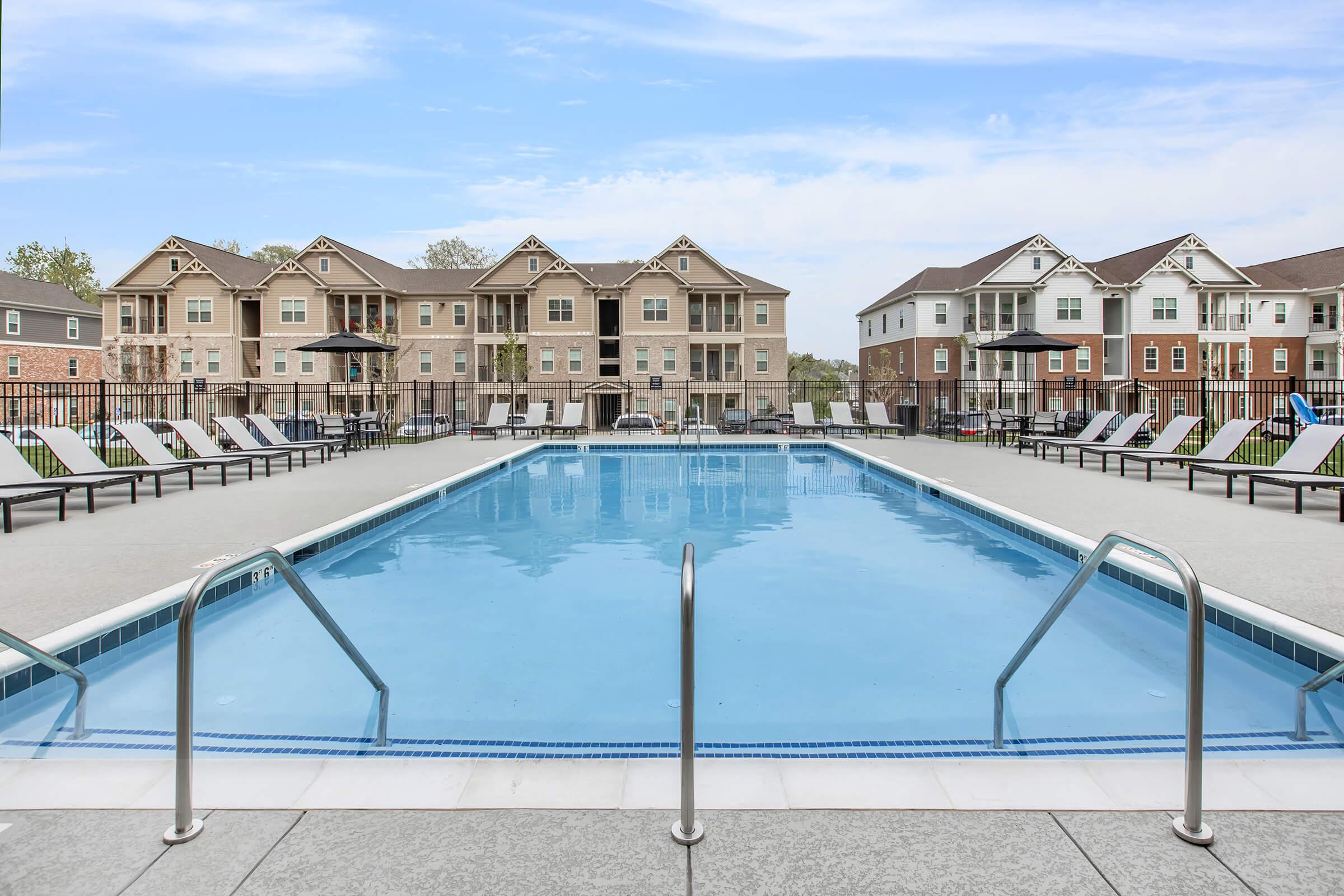 A clear swimming pool with metal handrails, surrounded by lounge chairs, and several multi-story residential buildings in the background. The sky is blue with some clouds, creating a bright and inviting atmosphere.