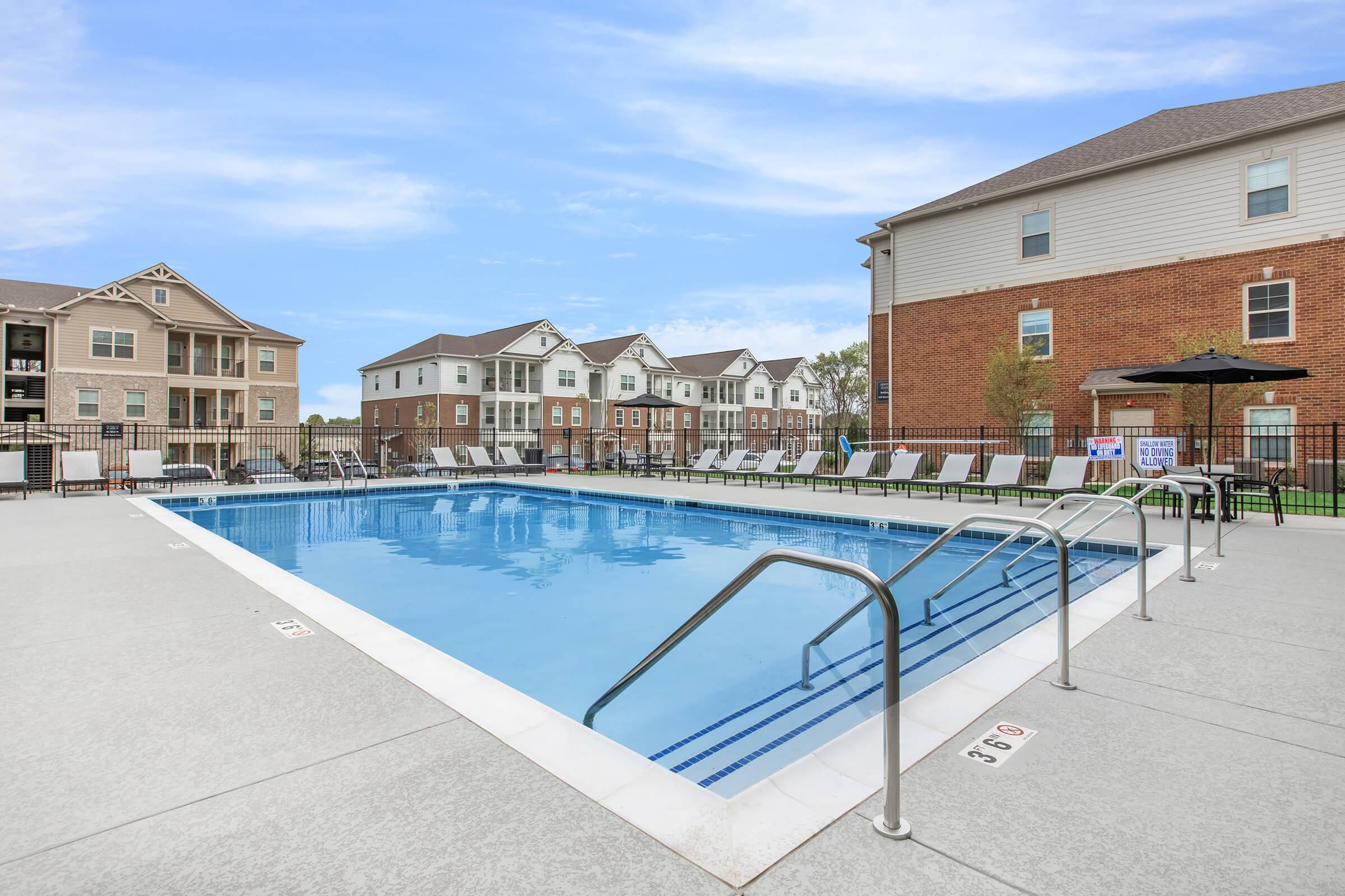 A clean, inviting swimming pool surrounded by lounge chairs, set against a backdrop of modern apartment buildings. The pool area features concrete decking and safety handrails, ideal for relaxation and recreation. Clear blue skies enhance the overall outdoor ambiance.