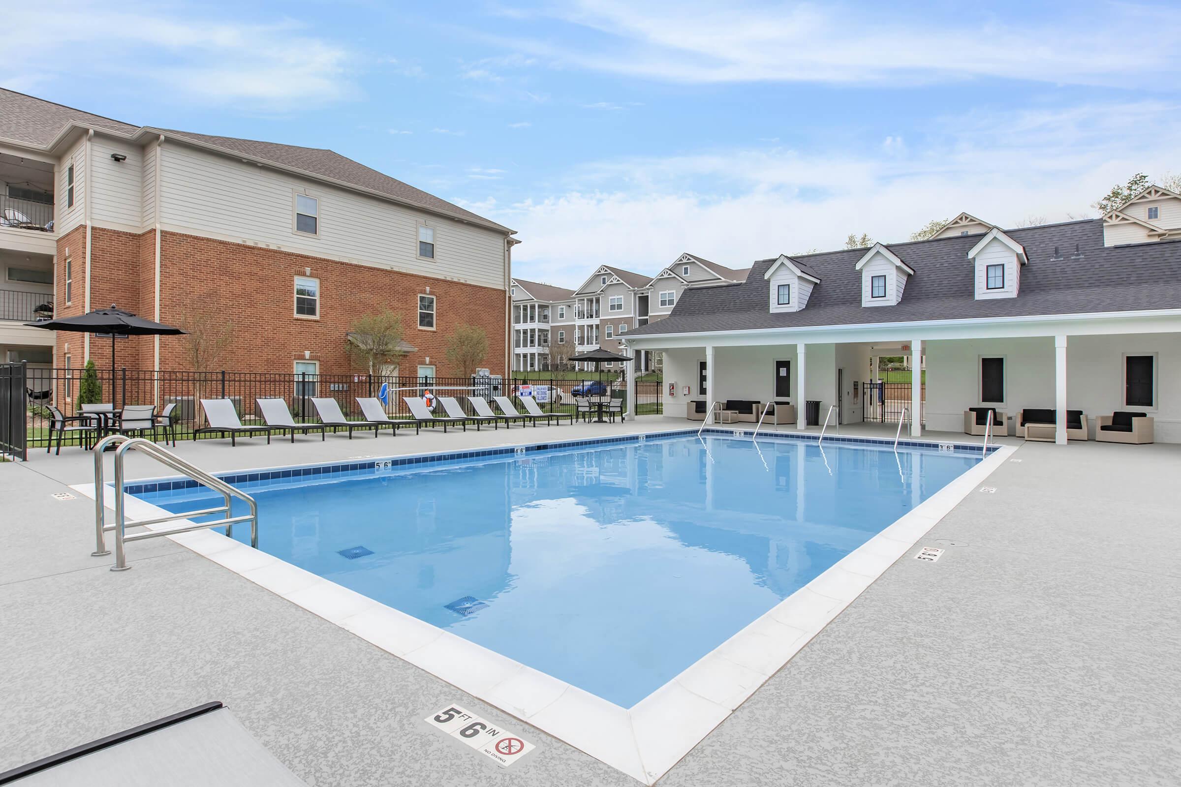 A clear swimming pool surrounded by lounge chairs and an outdoor building. The pool area features a safety railing and is enclosed by a fence. In the background, there are residential buildings with a mix of brick and siding. The sky is bright with a few clouds.