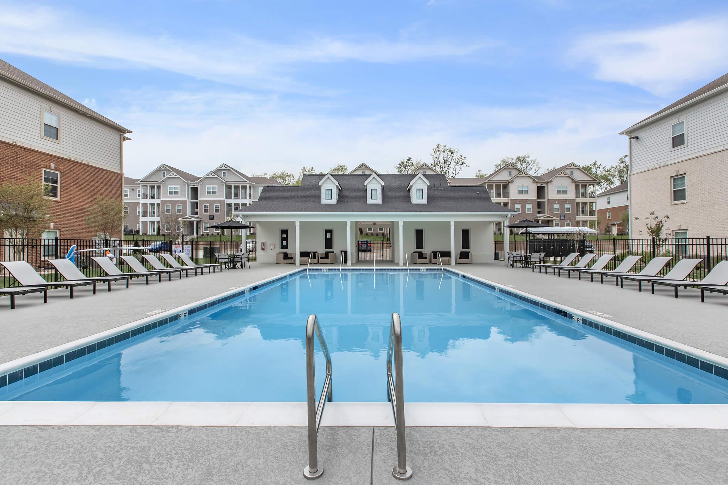 A clear blue swimming pool surrounded by lounge chairs, with a club house in the background. The area features landscaped greenery and several multi-story apartment buildings. The sky above is bright and partly cloudy, creating a inviting atmosphere for relaxation and leisure.