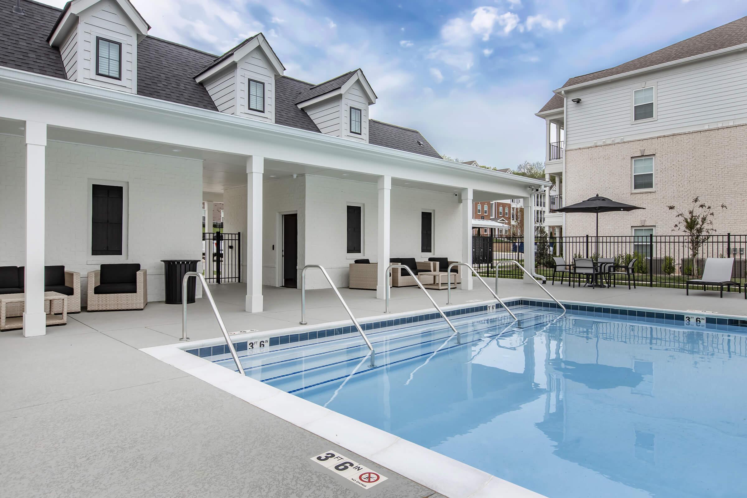A clear swimming pool with a gradual entry and handrails, situated beside a covered patio area featuring modern furniture. In the background, there are two buildings with a light exterior. The sky is partly cloudy, and the area is surrounded by a black fence and lush greenery.
