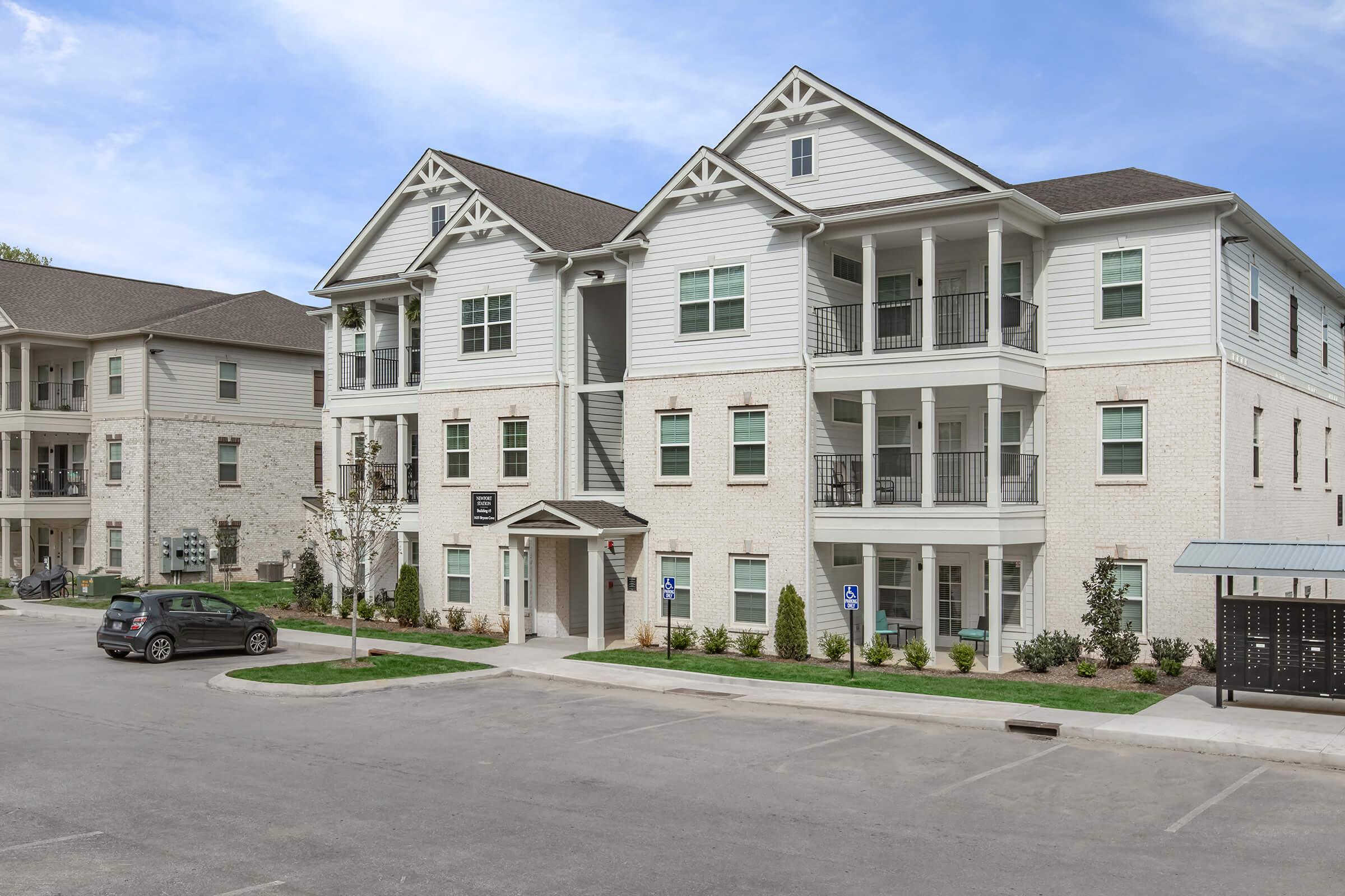 A modern three-story apartment building with a white exterior, featuring multiple balconies and large windows. Surrounding the building are landscaped areas with small bushes and trees, along with an empty parking lot. The sky above is clear with a hint of blue.