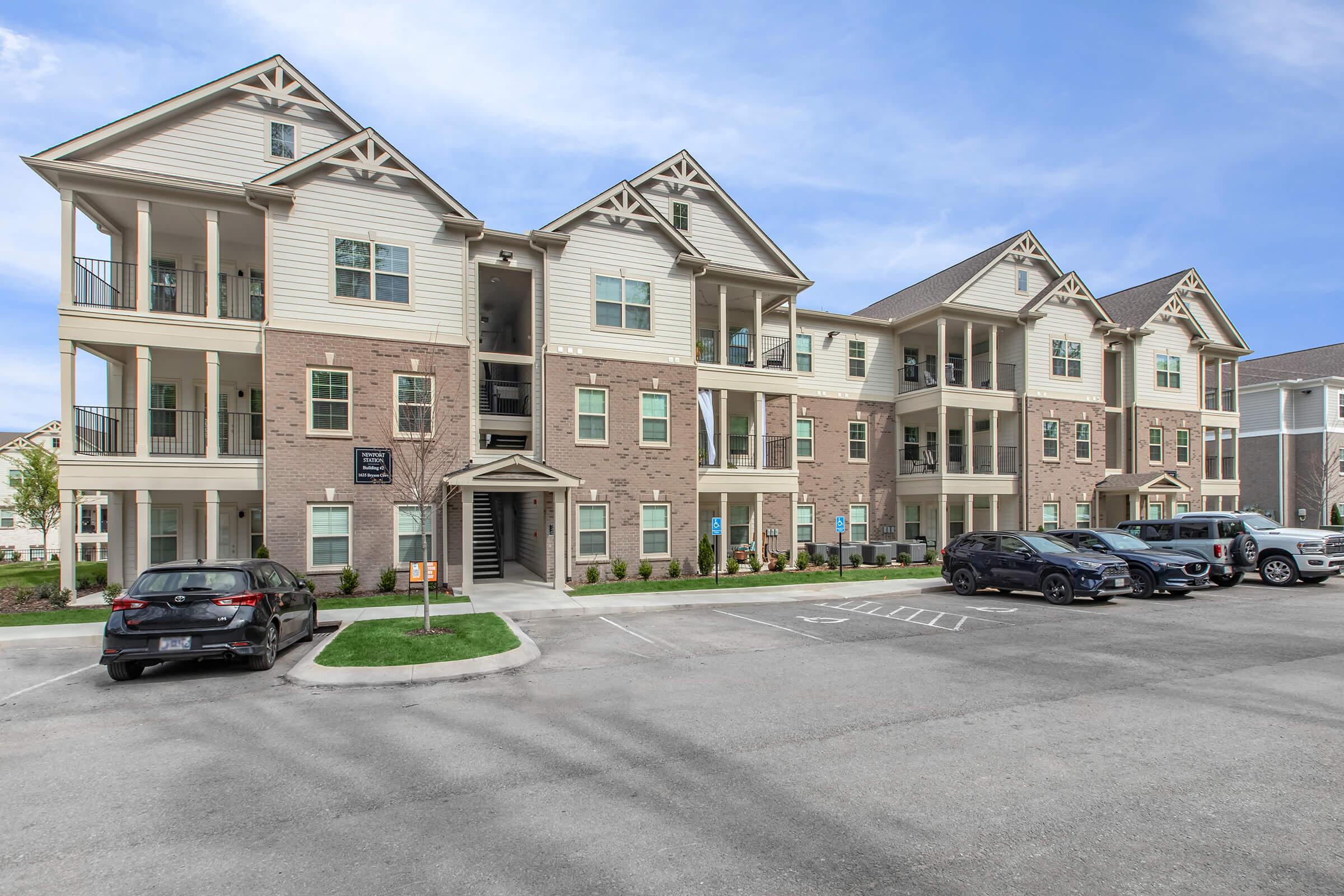 A multistory apartment building featuring a combination of brick and siding exteriors. Multiple balconies are visible on the upper floors. The parking area in front includes several parked cars. The sky is clear with a few clouds, and there are landscaped areas around the building.
