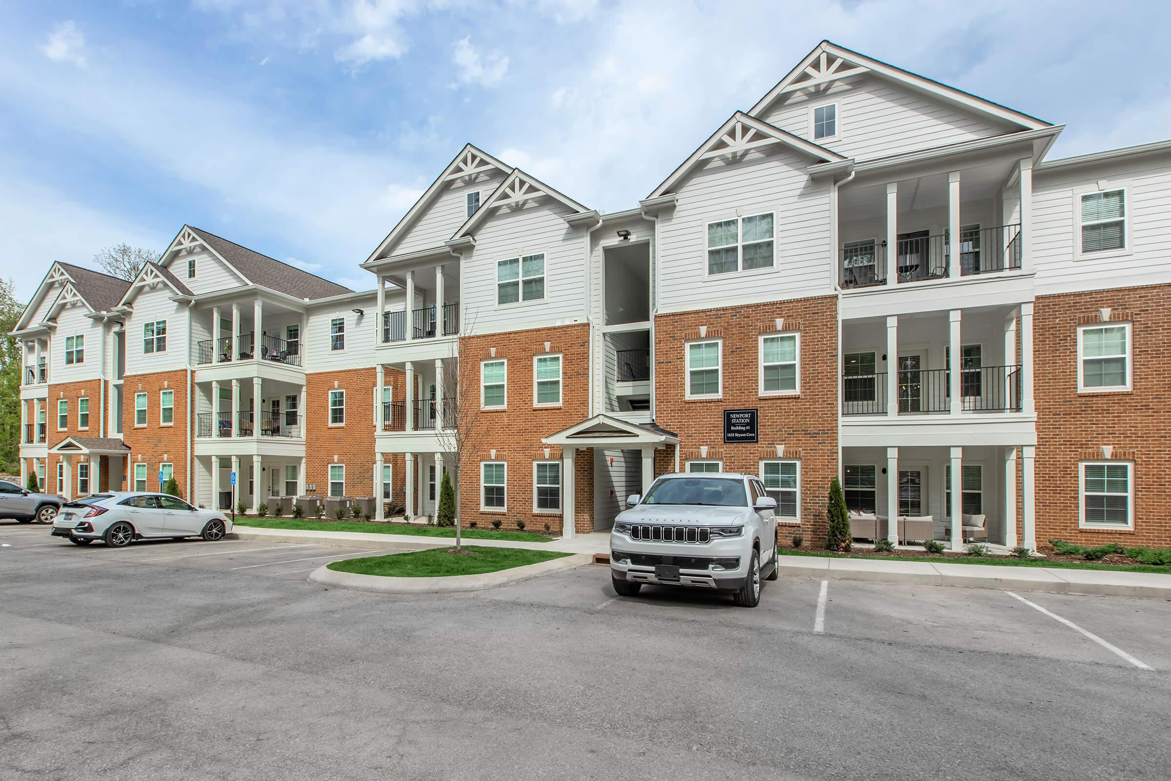 A multi-story apartment building featuring a mix of brick and white siding. The facade includes multiple balconies, large windows, and a welcoming entrance. There is a parking area in front with a few parked cars, set against a clear sky. Green landscaping borders the building.