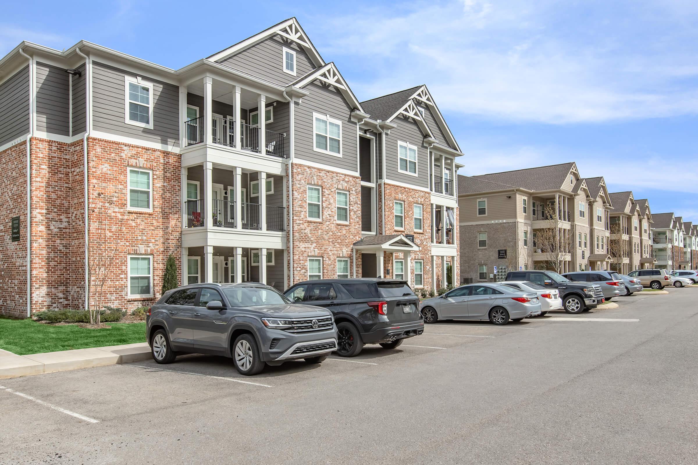 A view of a modern residential apartment complex featuring brick and gray siding. The building has multiple balconies and is surrounded by a parking lot filled with various cars. Clear blue skies and green lawns add to the aesthetic of the neighborhood.