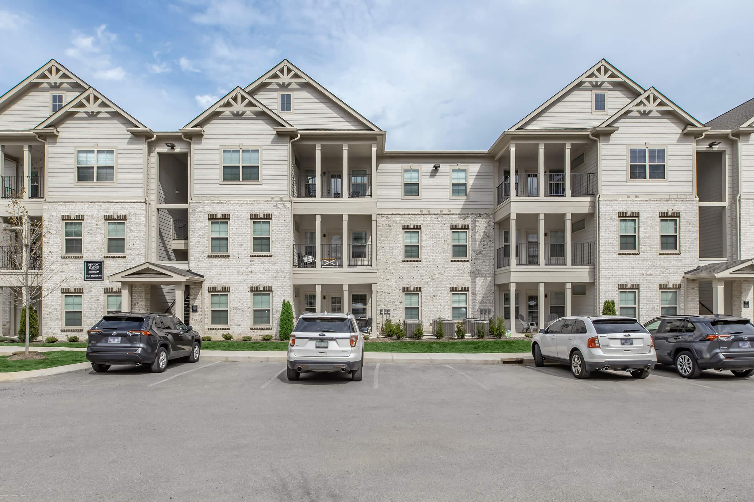 A three-story apartment building with a light-colored exterior featuring a mix of brick and siding. The structure has multiple balconies and a row of parked cars in the foreground. The sky is partly cloudy, and there is landscaped greenery around the building.