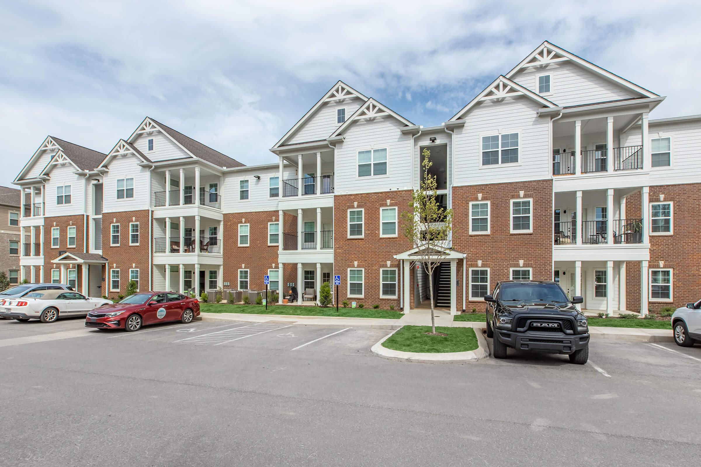 A modern multi-unit apartment building with a mix of brick and white siding exteriors. The building features multiple balconies, decorative peaked roofs, and a covered entrance. Several parked cars are visible in the foreground, along with landscaped greenery.