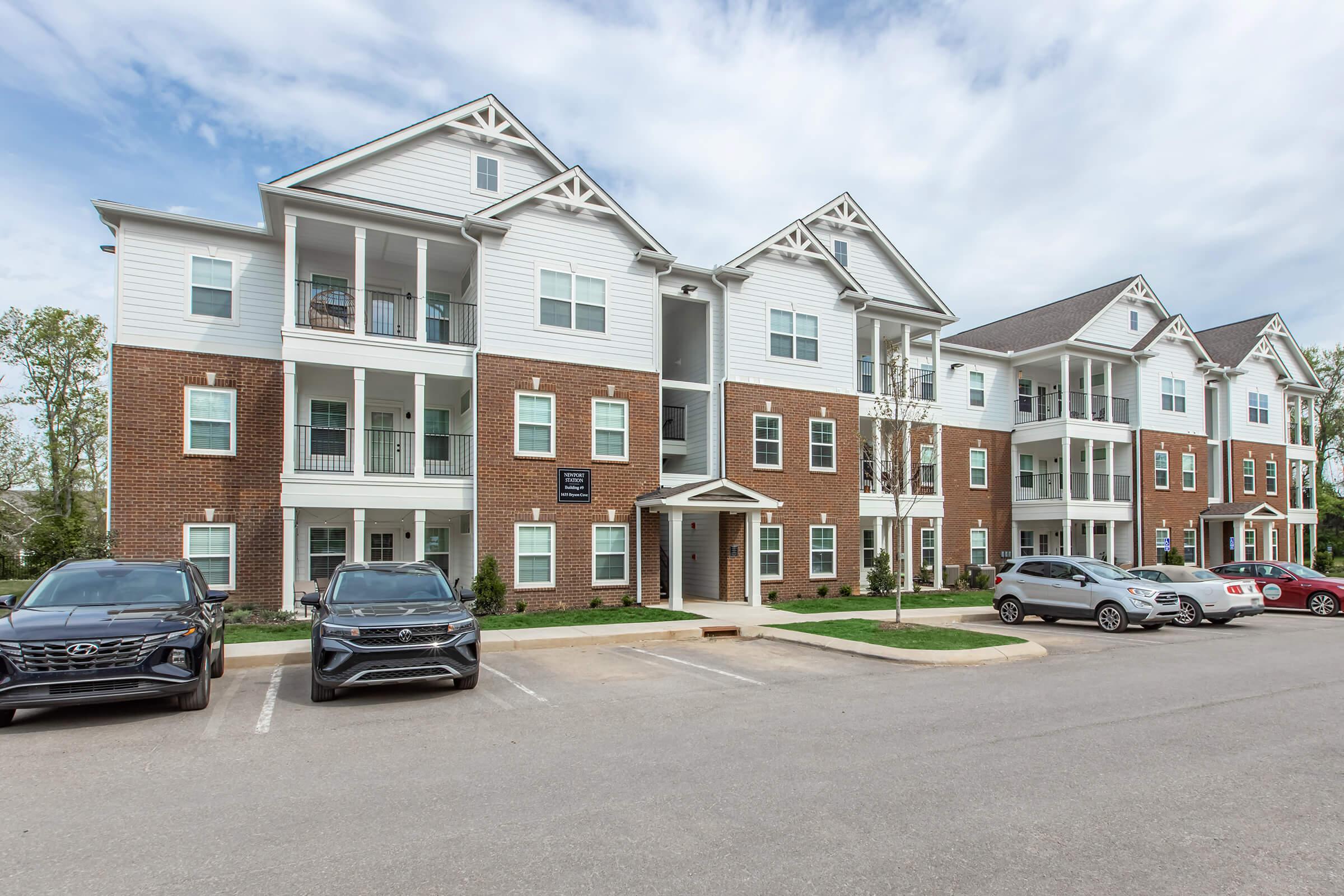 Modern apartment building featuring a mix of brick and siding exterior with multiple stories, balconies, and large windows. A parking lot is visible in front with several cars parked. The sky is partly cloudy, and there are manicured lawns and greenery around the building.