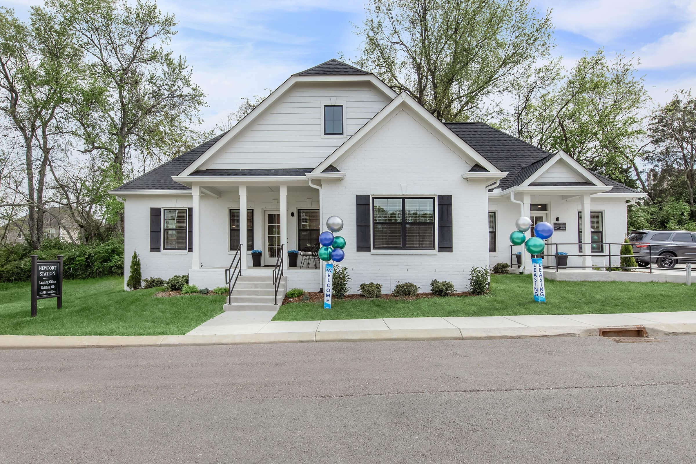 A modern, single-story white house with a black roof, featuring a porch with black railings. Two decorative columns with blue and green balloons are placed at the entrance. Green grass and small shrubs surround the house, and a paved walkway leads to the front door. The street is visible in the foreground.