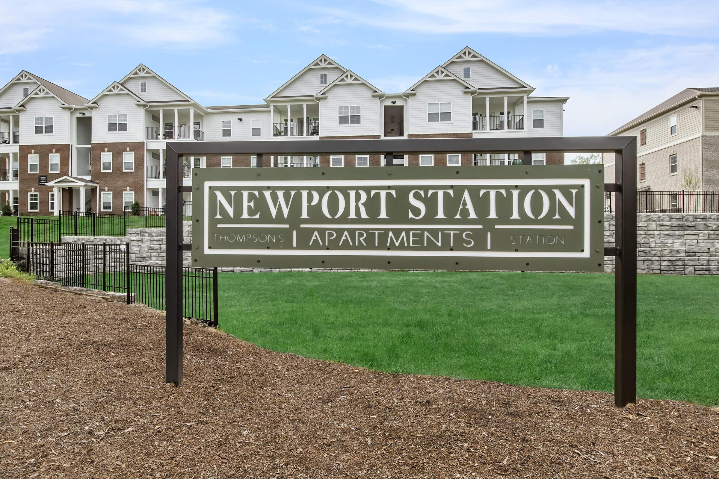 Sign for Newport Station Apartments, featuring the name prominently displayed. The background includes a well-maintained lawn and modern apartment buildings, emphasizing a residential area. The sign is mounted on a decorative frame, indicating a welcoming entrance to the apartment complex.
