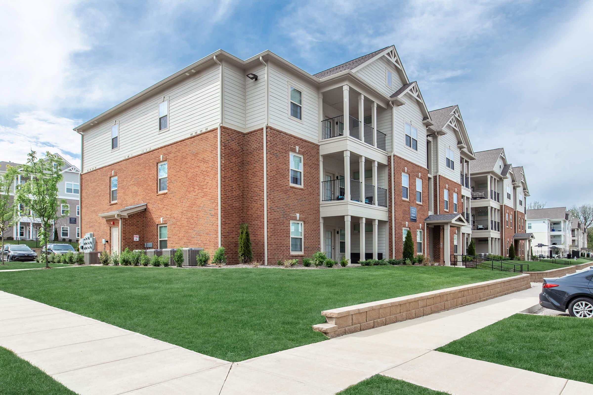A modern apartment building with a mix of brick and siding, featuring multiple balconies, landscaped green lawns, and a paved walkway in the foreground. The sky is partly cloudy, and there are cars parked nearby, creating a welcoming residential neighborhood atmosphere.