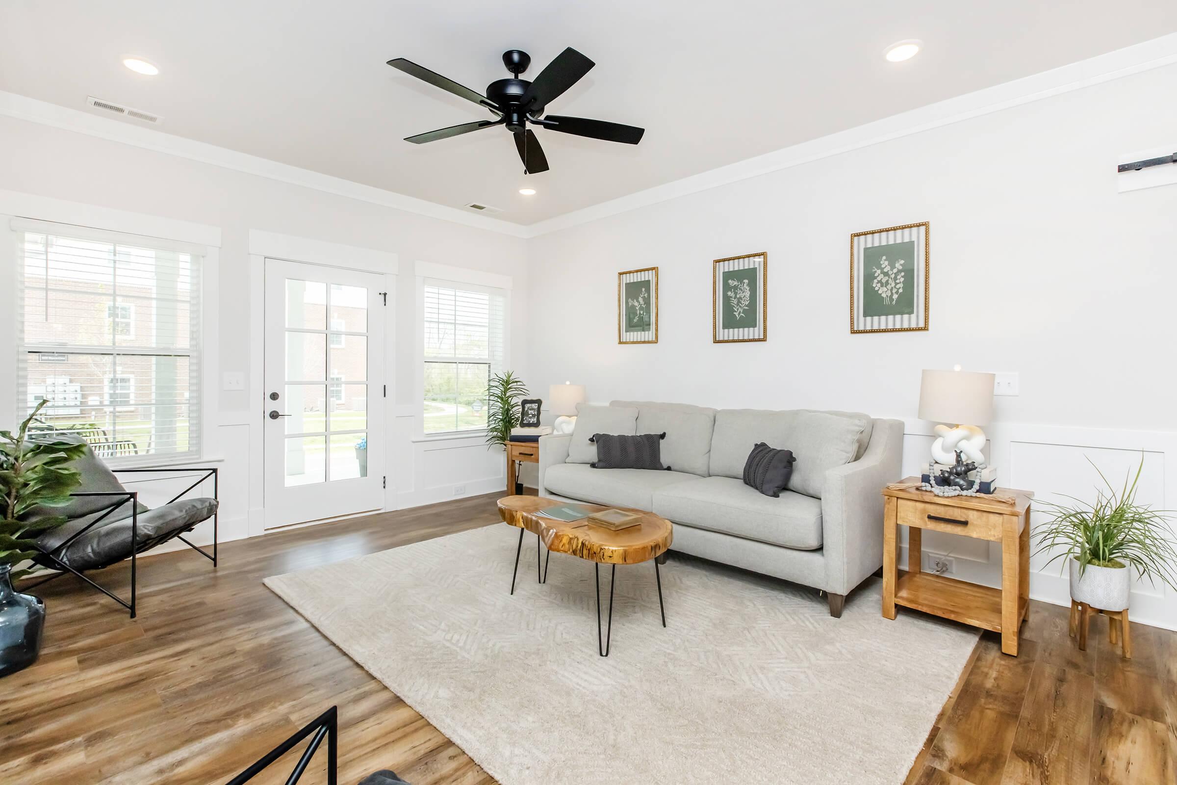 A bright and airy living room featuring a light gray sofa, a wooden coffee table, and a decorative rug. There are large windows allowing natural light, potted plants for greenery, and framed wall art. A ceiling fan and stylish furniture complete the cozy atmosphere.