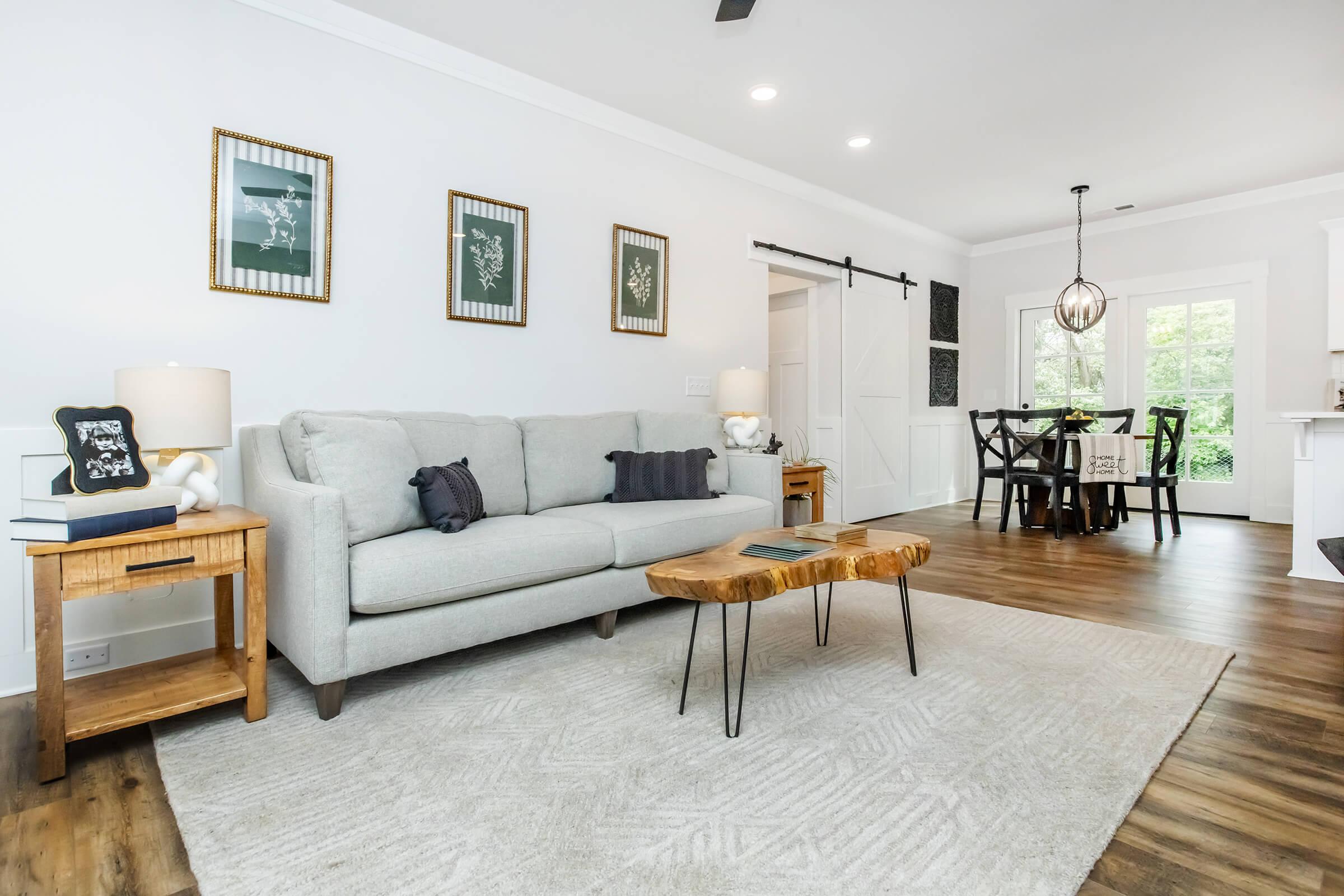 A cozy living room featuring a light gray sofa with dark cushions, a natural wood coffee table, and two side tables with lamps. The space is adorned with framed botanical art on the walls, a plush area rug, and an open dining area with a wooden table and chairs. Large windows allow natural light to fill the room.