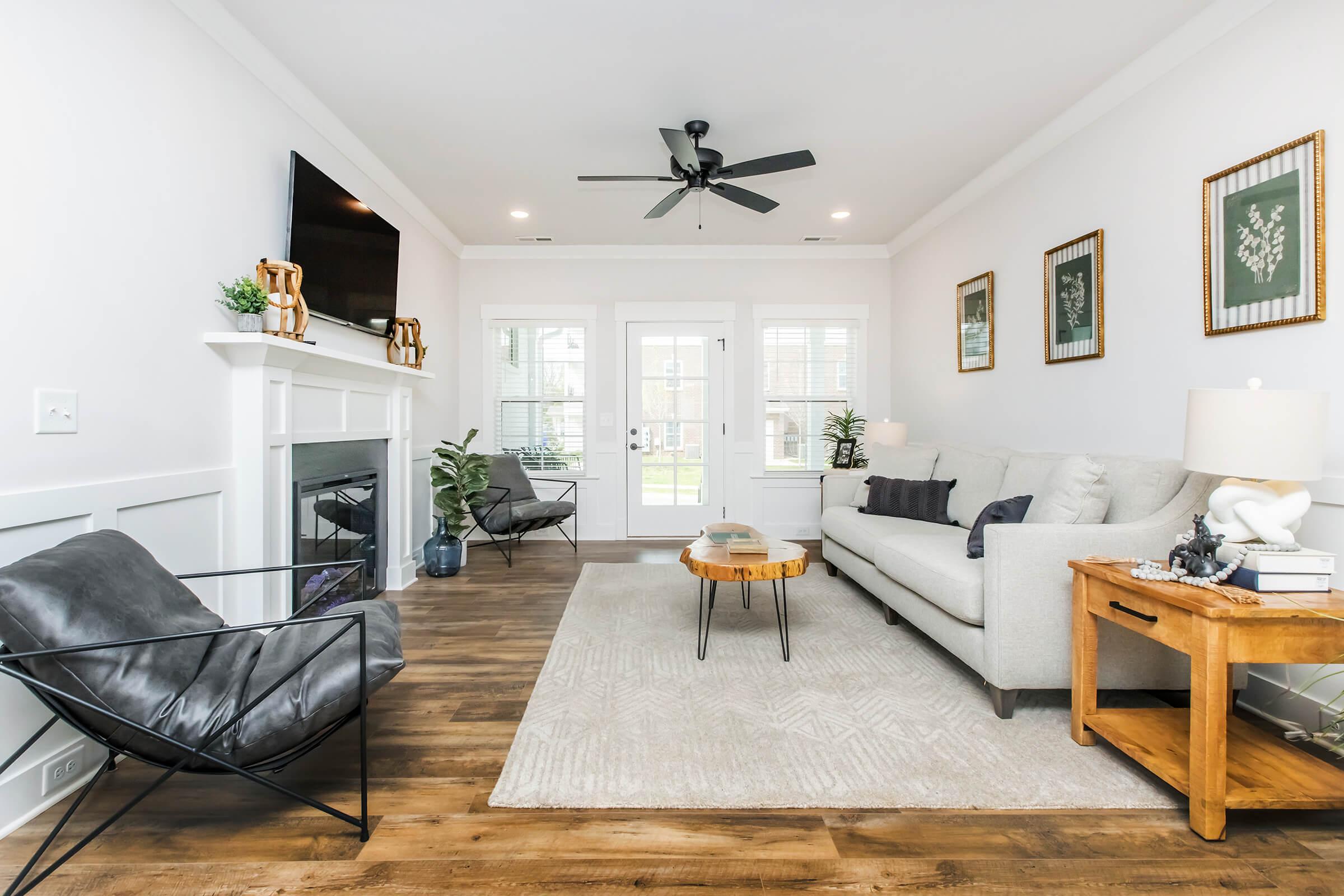 A bright and modern living room featuring a gray sofa, a wooden coffee table, and a black accent chair. The decor includes framed botanical prints on the walls and a ceiling fan above. Large windows allow natural light to enter, creating a welcoming atmosphere. The flooring is wooden, enhancing the room's warmth.