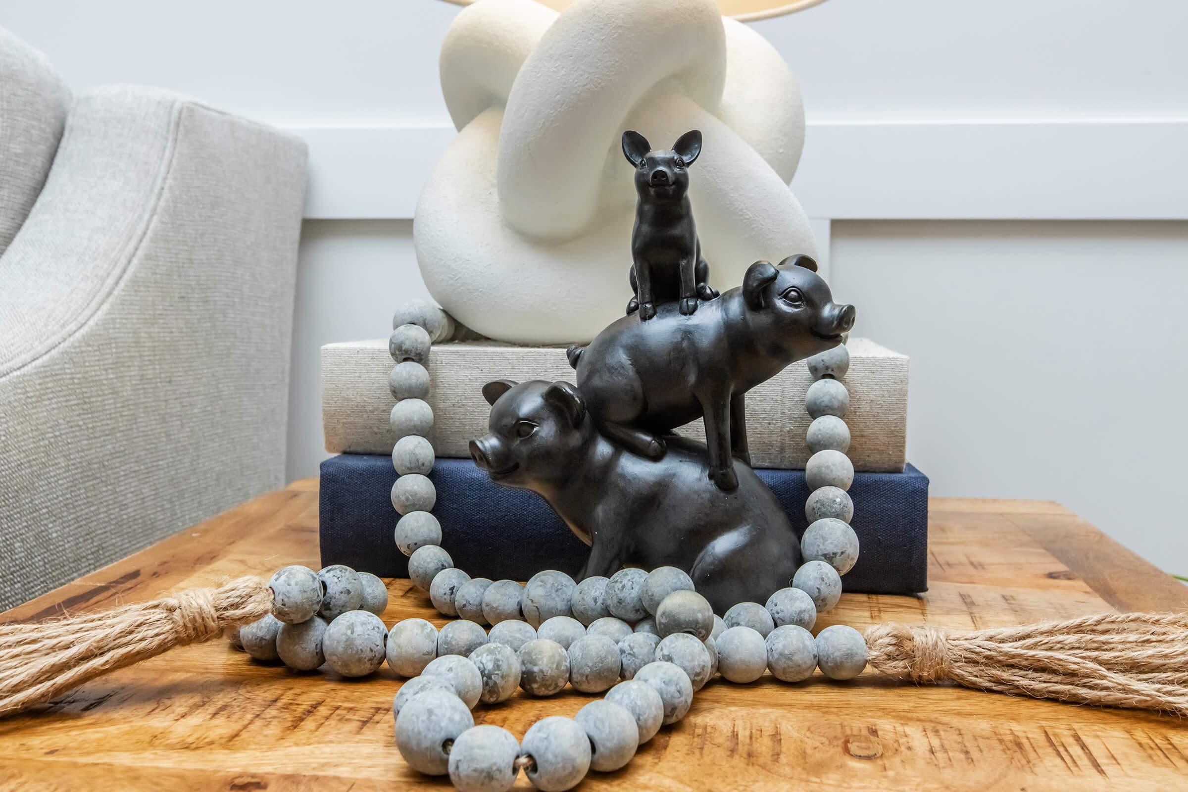 A decorative arrangement featuring three black animal figurines—a small dog on top of two pigs—resting on a stack of books. A textured cream sculpture and a beaded garland with tassels made of gray beads are included in the composition, all displayed on a wooden table.