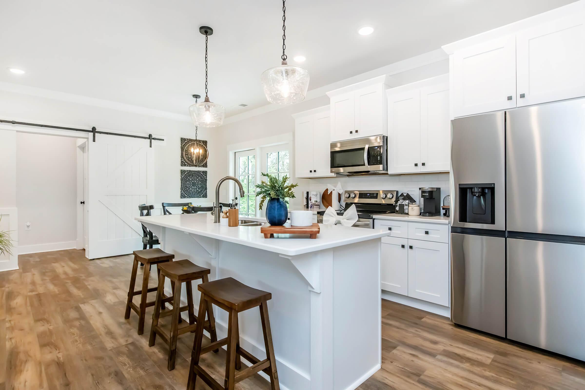 Modern kitchen featuring a large island with three wooden bar stools, stainless steel appliances, and white cabinetry. Pendant lights hang above the island, and a door with a sliding barn style is visible in the background. The space is bright and spacious with natural light coming in through windows.