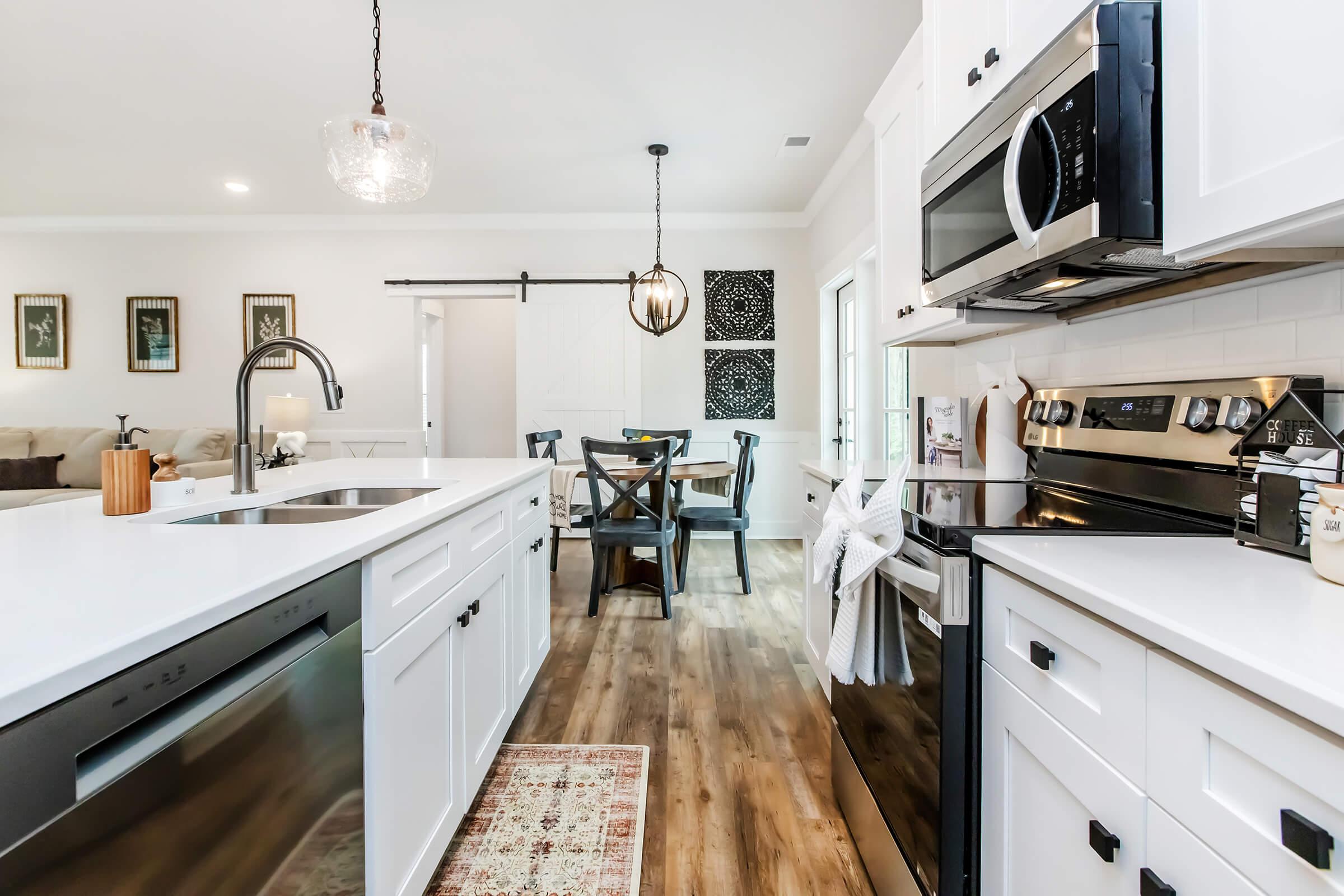 A modern kitchen featuring white cabinets, stainless steel appliances, and a central island with a sink. In the background, a dining area with a wooden table and chairs is visible, along with decorative wall art and a sliding barn door. The flooring is a warm wood finish, creating a cozy atmosphere.