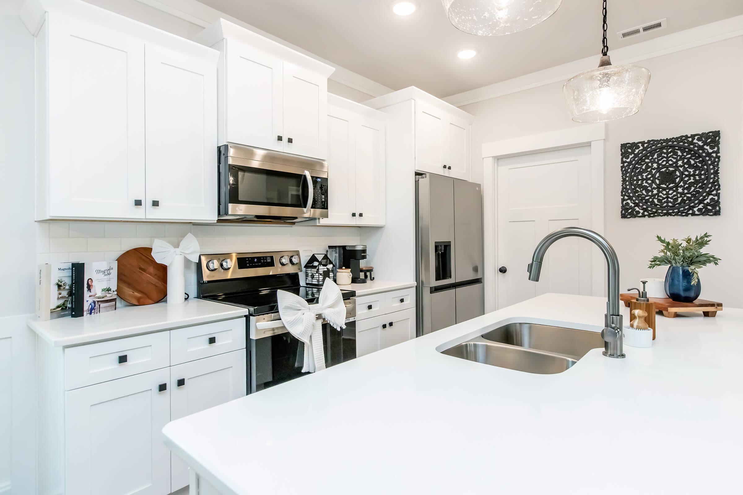 A modern kitchen featuring white cabinets, stainless steel appliances, and a large island with a sink. The countertop is bright and clean, and decorative elements include a vase with flowers and a wooden board. The overall design is sleek and inviting, with soft lighting and contemporary decor.