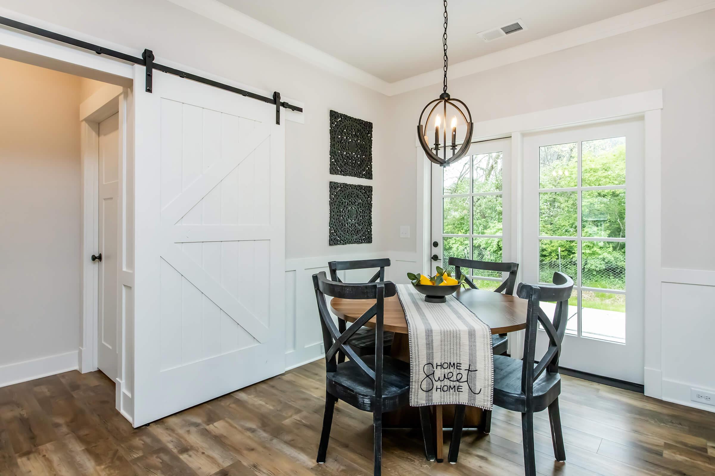 A modern dining area featuring a round wooden table surrounded by black chairs. A decorative table runner with the words "HOME SWEET HOME" is centered on the table. Nearby, a sliding barn door and a large window with multiple panes provide natural light, with greenery visible outside.