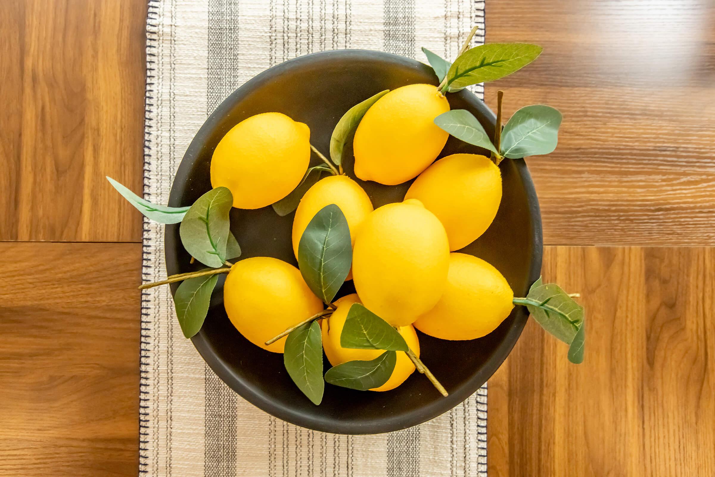 A round black bowl filled with bright yellow lemons, accented with green leaves, placed on a striped table runner. The background features a wooden surface, creating a warm and inviting atmosphere.